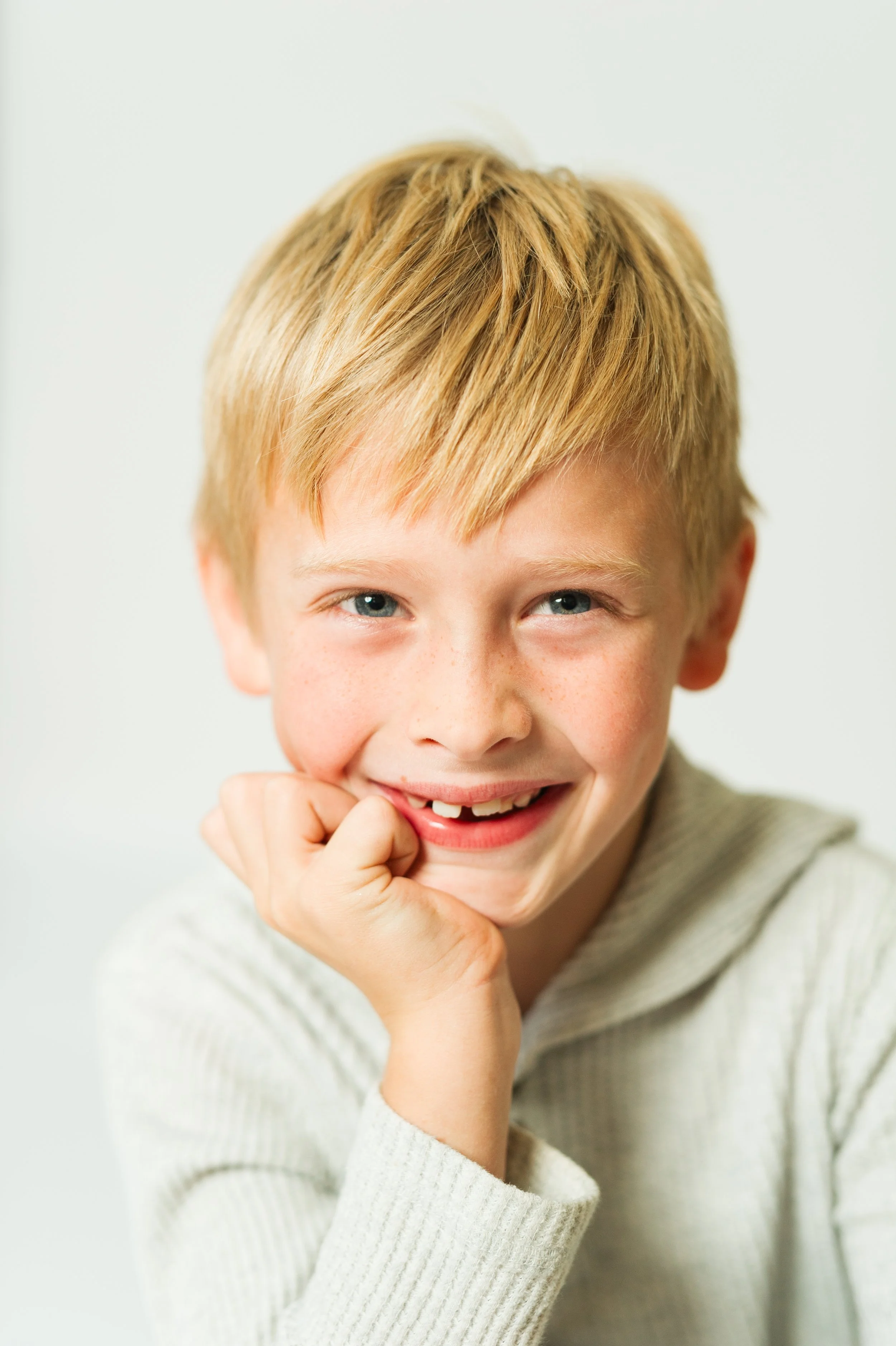 Smiling child with blond hair and missing teeth, wearing a light gray sweater, resting chin on hand.