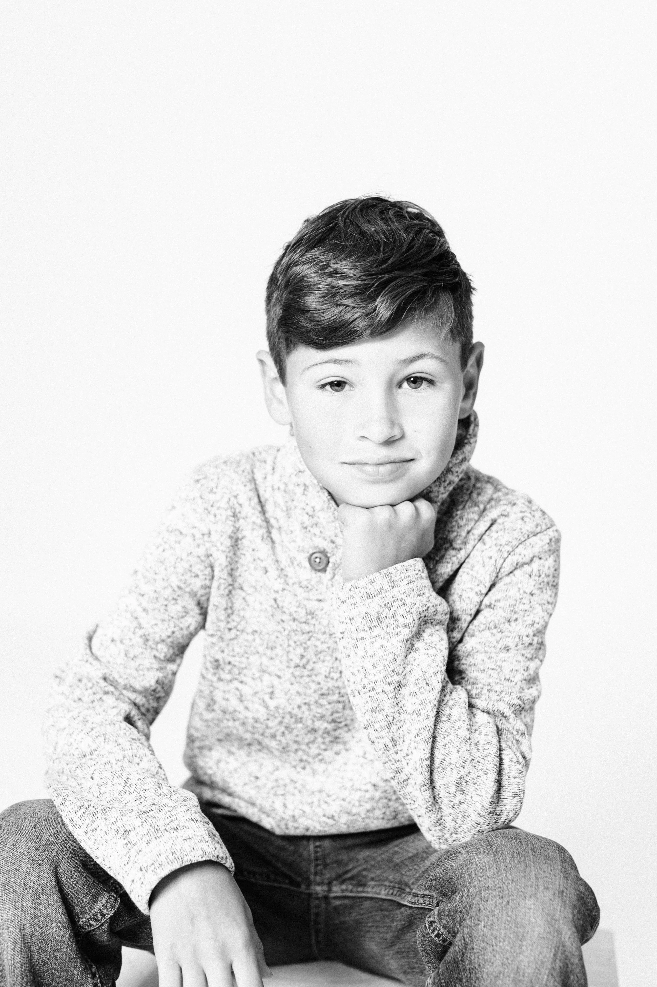 Black and white portrait of a boy wearing a sweater and jeans, resting his chin on his hand.