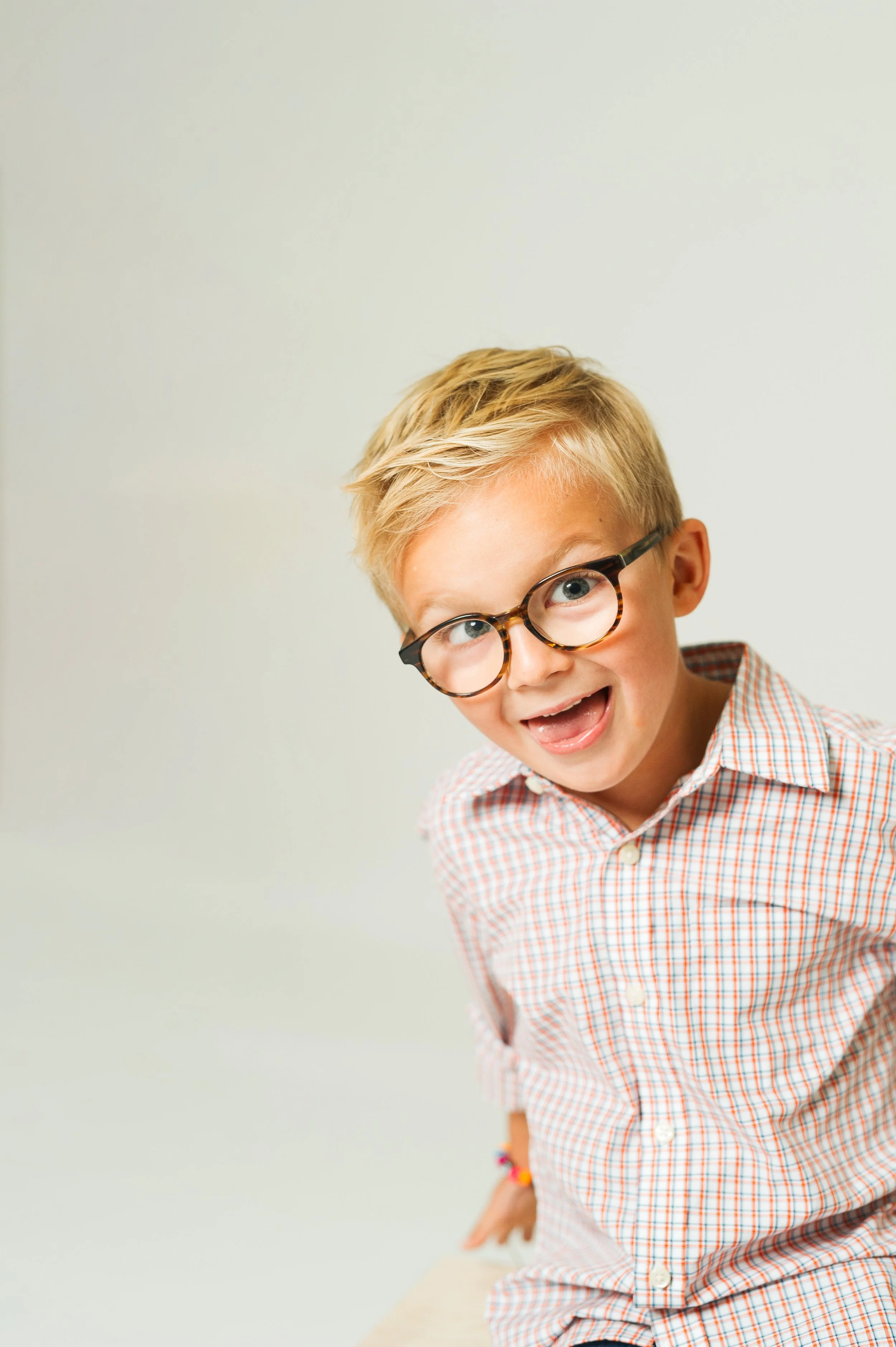 Young boy with blond hair wearing glasses and a plaid shirt, smiling playfully.