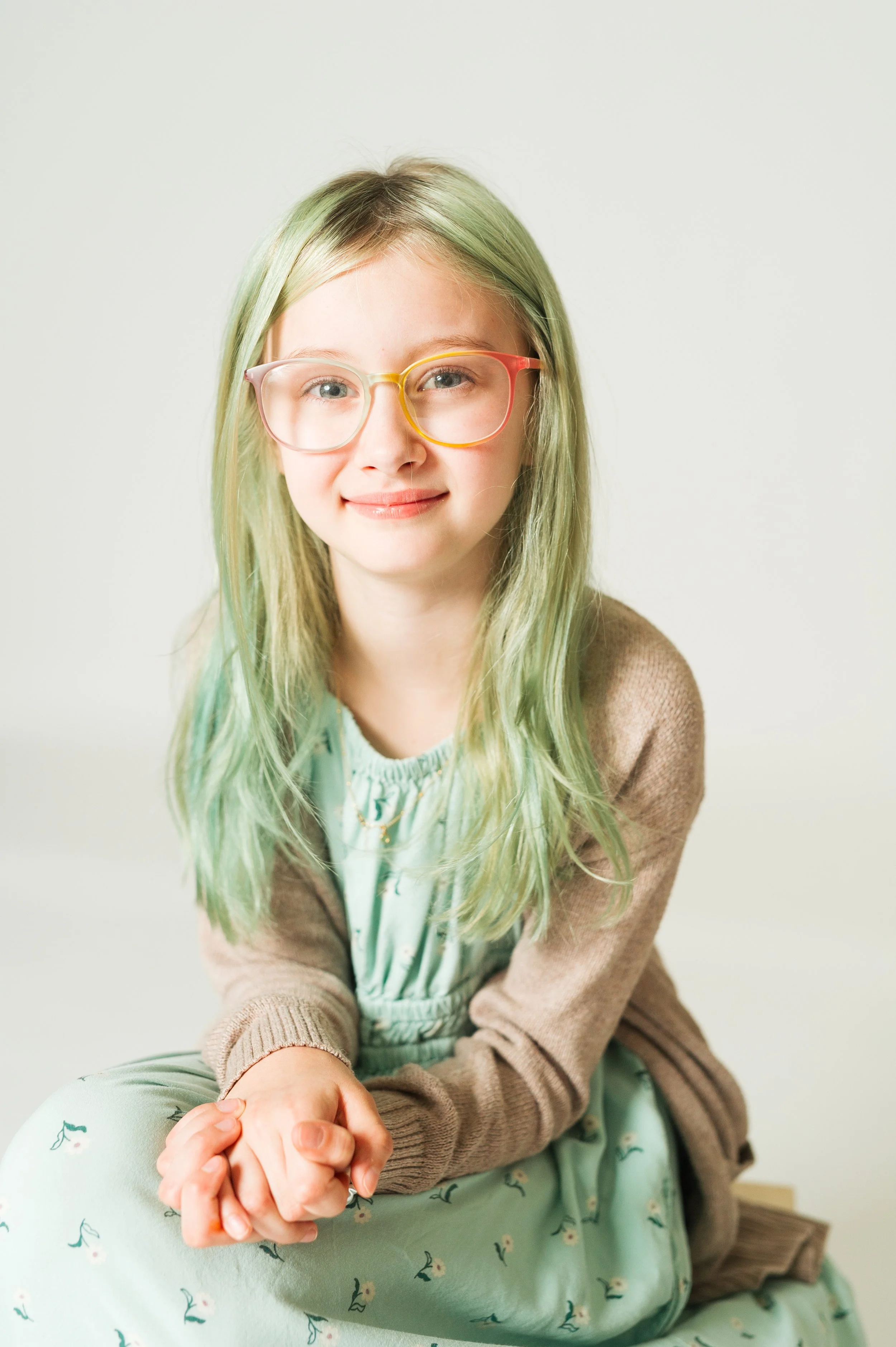 Young girl with green hair and glasses, wearing a light green dress with floral patterns and a beige cardigan, sitting and smiling in a studio setting.