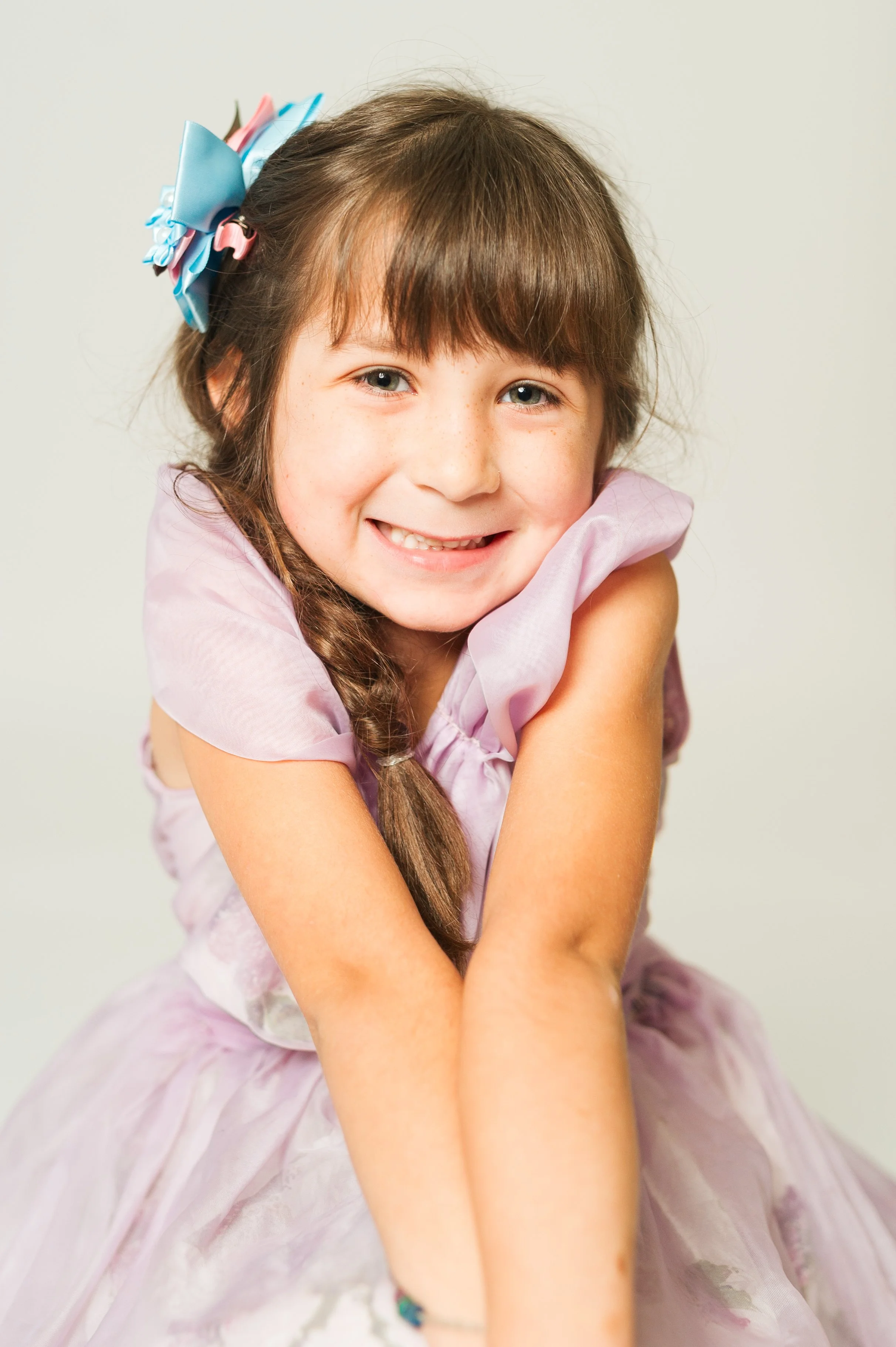 Young girl in a purple dress, smiling with arms clasped, wearing a blue hair bow.