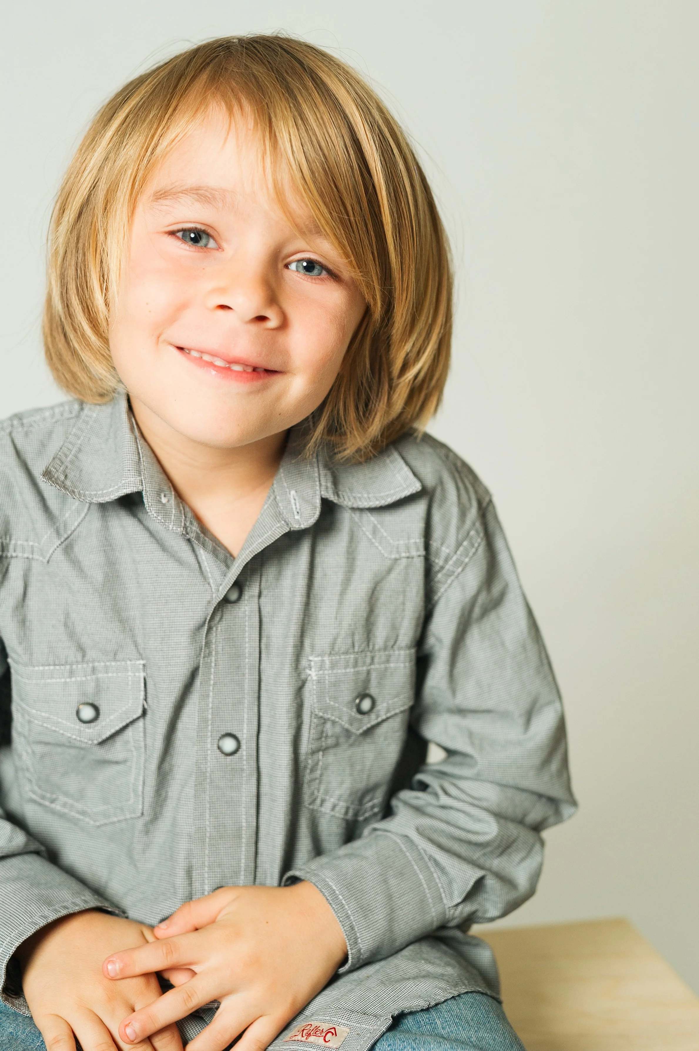 Young boy smiling, wearing a gray button-up shirt, sitting indoors against a plain background.