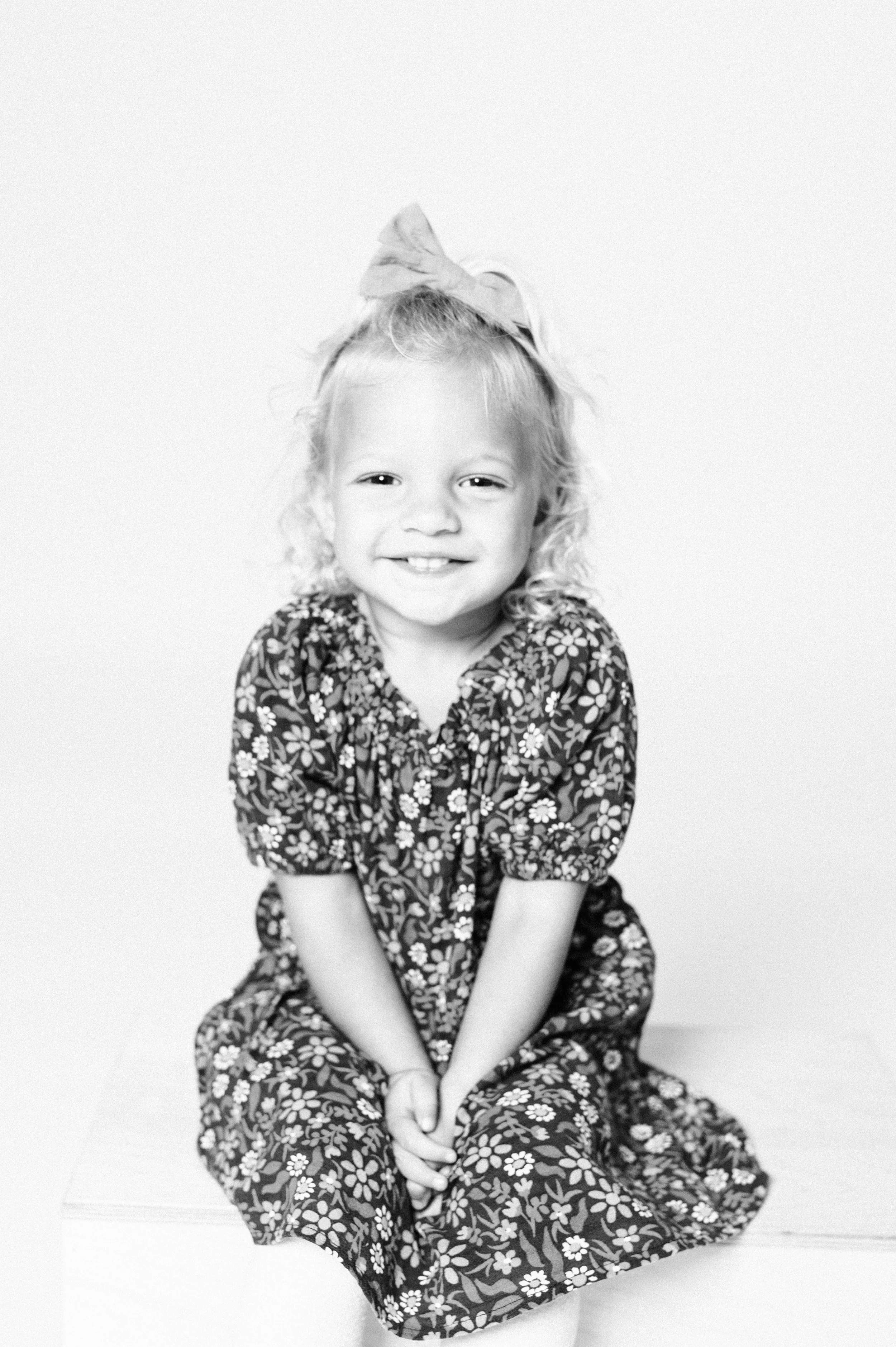 Smiling child in floral dress and bow sitting on a stool, black and white photo.