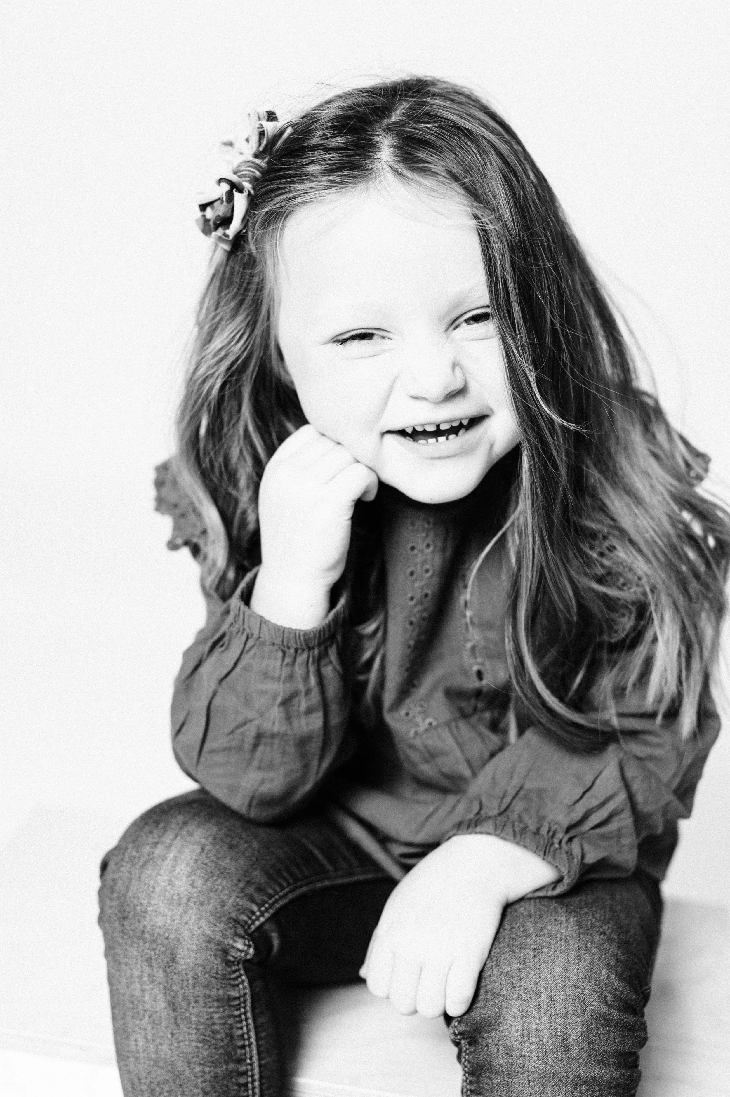 Young child with long hair, smiling, wearing a dark shirt and jeans, black and white portrait.