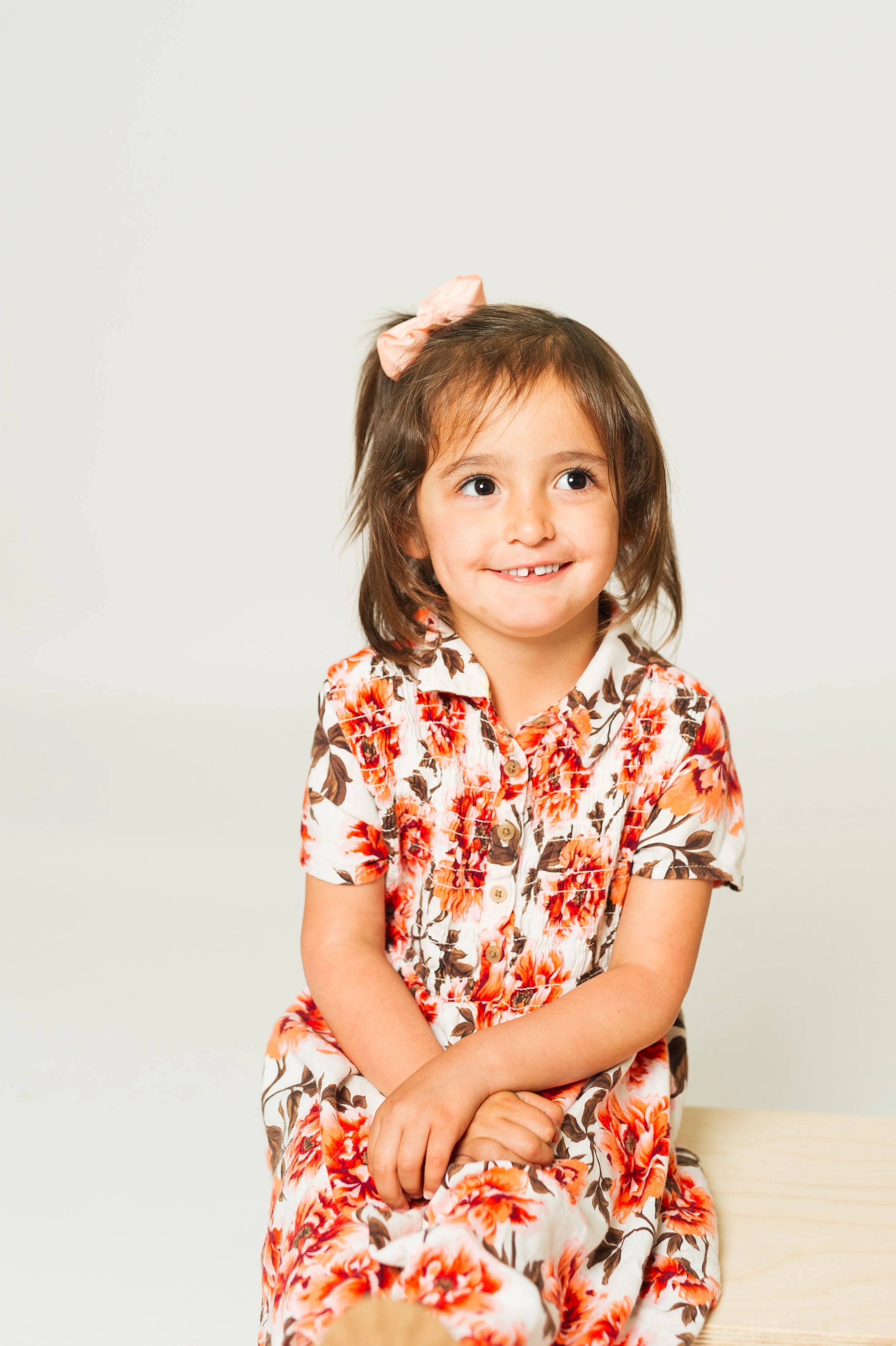A young girl wearing a floral dress with a bow in her hair, sitting and smiling against a plain background.