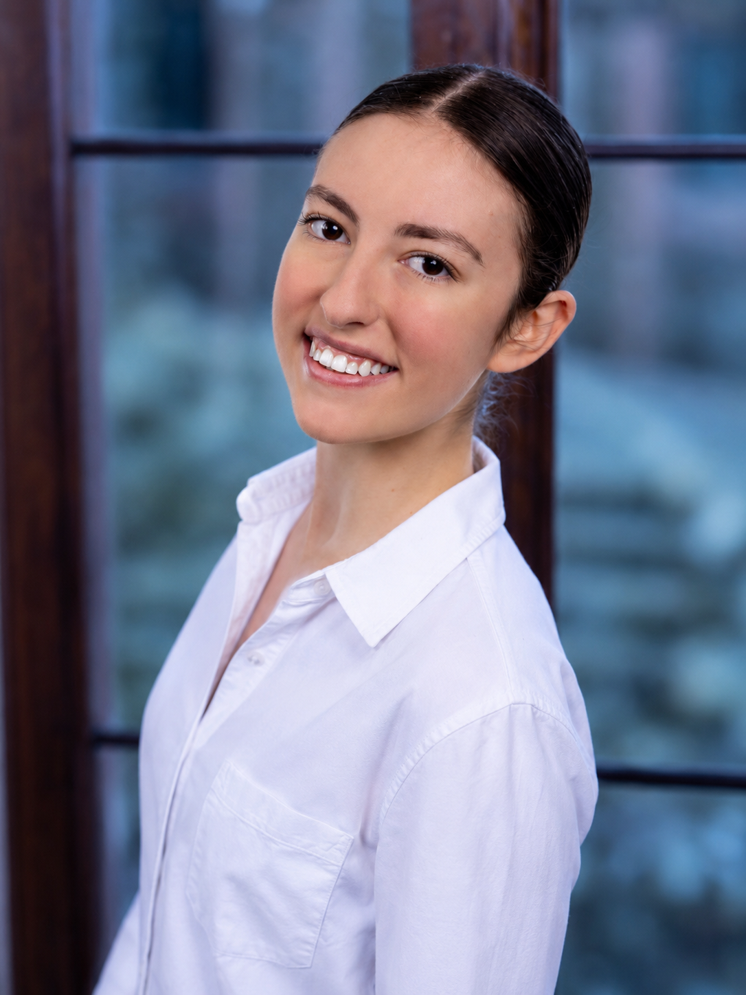 A young woman with long brown hair, wearing a white shirt, holding her hand near her face, looking directly at the camera, in front of a window with a blurred outdoor background.