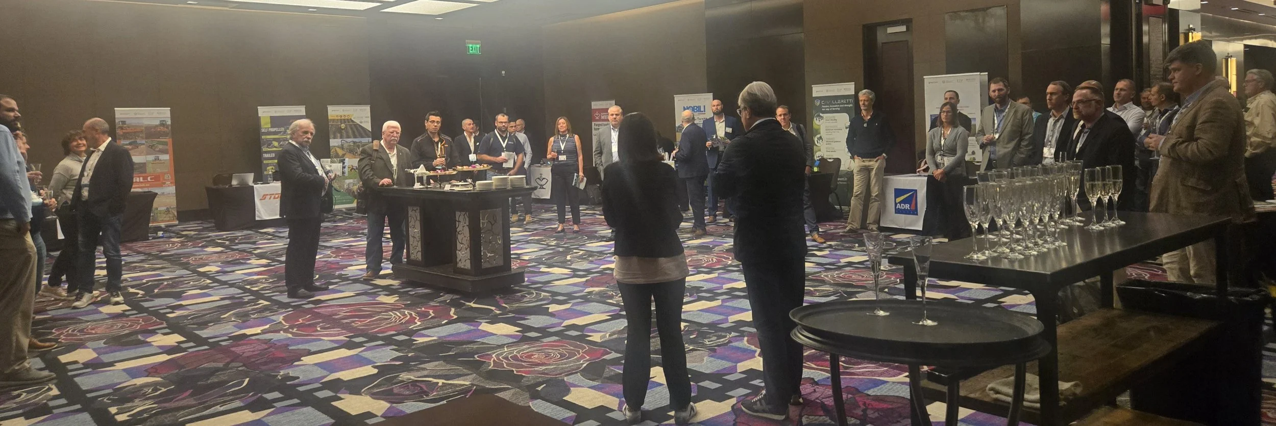 A group of people attending a professional conference or event in a large, carpeted room with display booths in the background. Some individuals are engaged in conversation, while others are standing near tables with food and drinks.