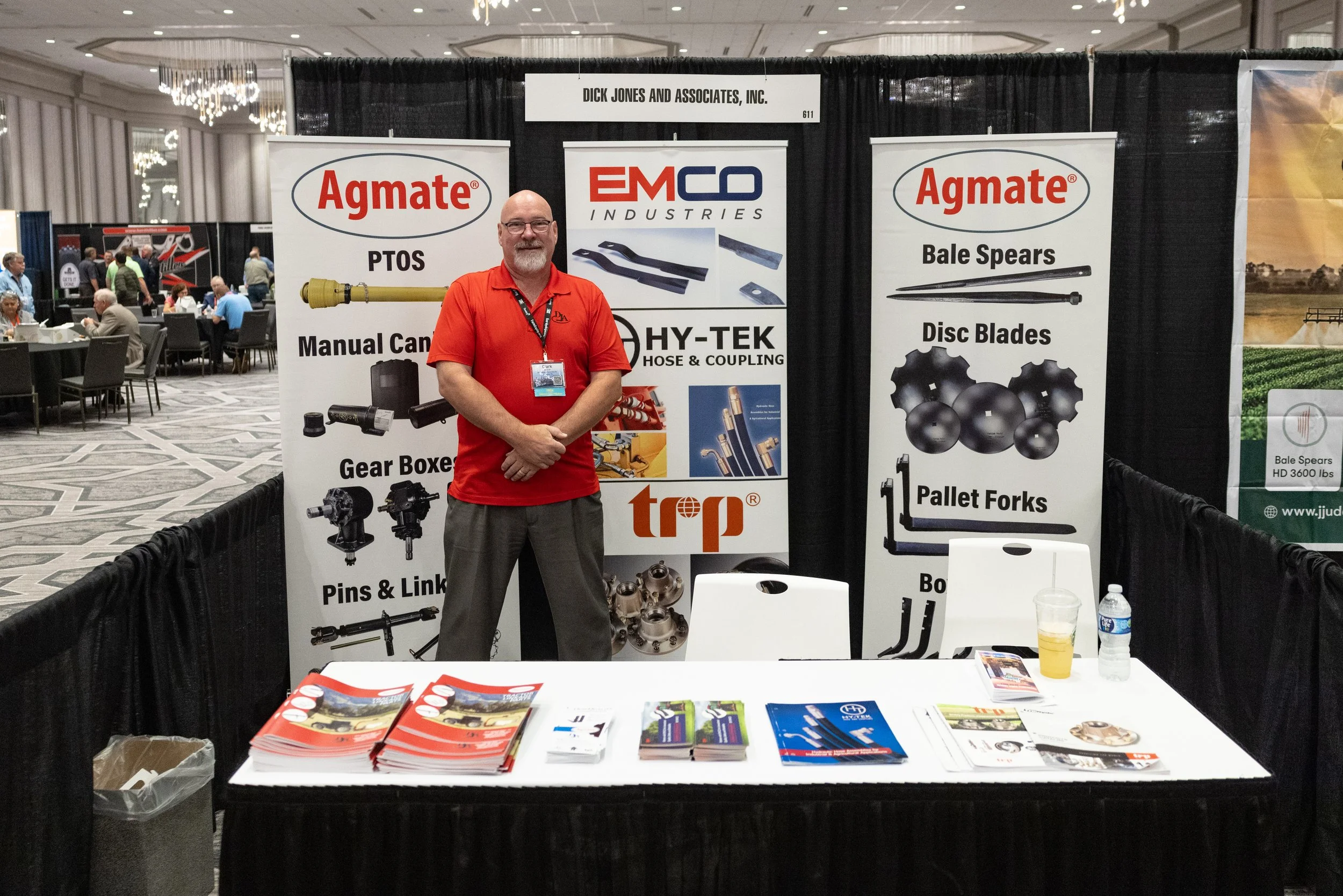 Man standing at a trade show booth with banners displaying industrial tools and components, including PTOS, manual cam, gear boxes, pins, links, bale spears, disc blades, and pallet forks. The booth has brochures and promotional materials on the table.