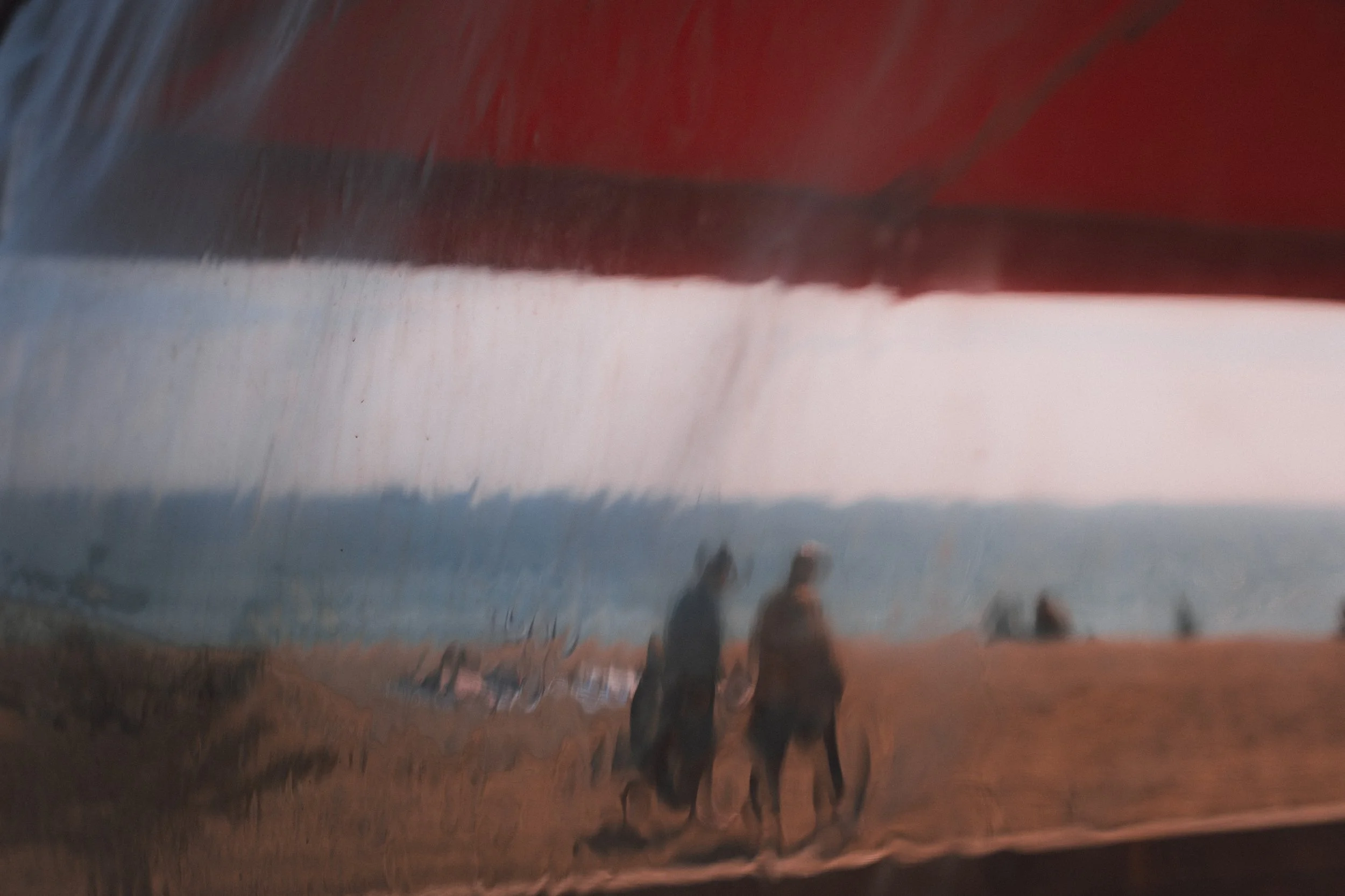 Reflected image of two people walking on a beach with a boat in the distance during sunset or dawn, viewed through a window with streaks of water