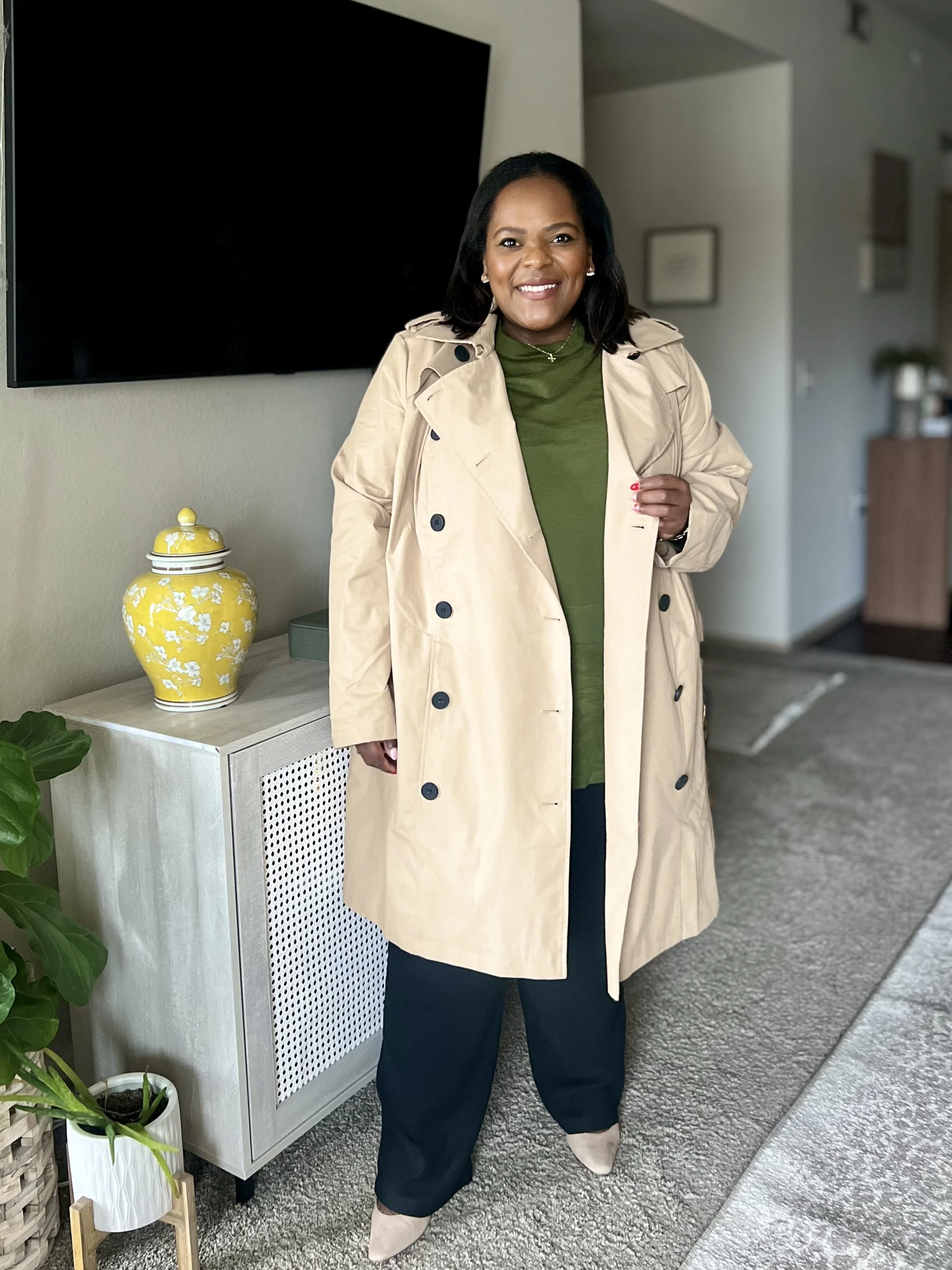Person in a beige trench coat and green top standing indoors near decor items.
