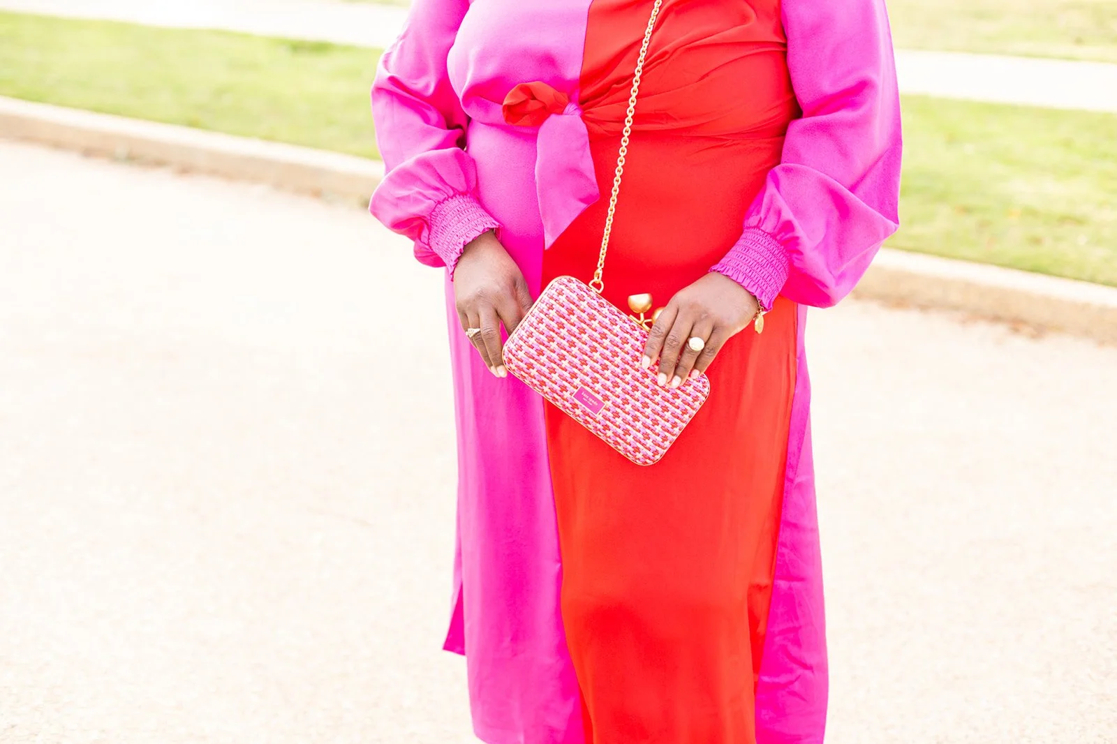 Person wearing a pink and red outfit holding a patterned clutch purse with a gold chain.