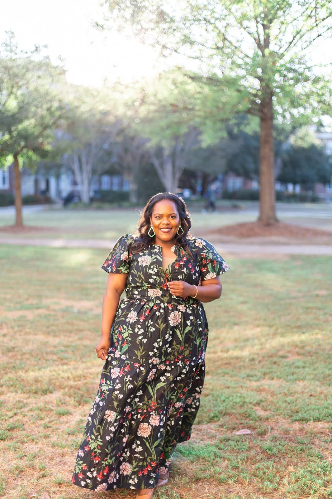 Woman in floral dress walking in a park with trees and sunlight in the background.