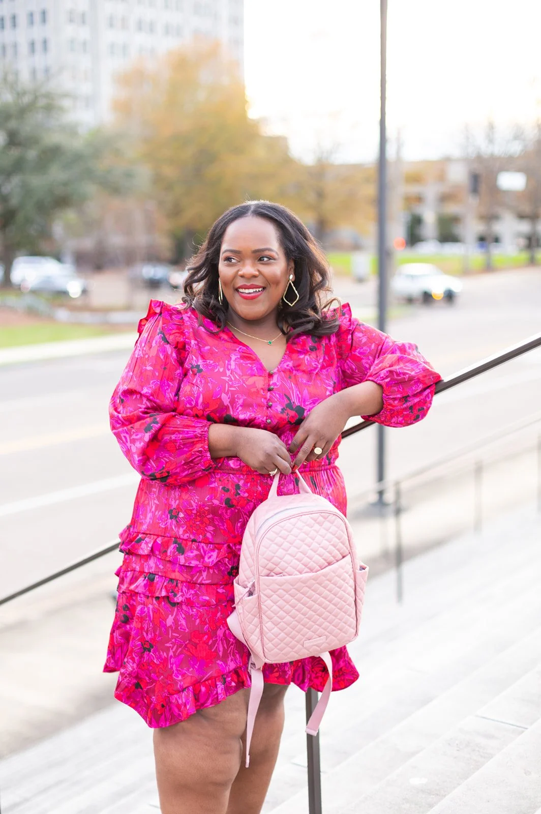 Woman in a pink floral dress holding a quilted pink backpack, standing outdoors on steps with blurred background of trees and buildings.