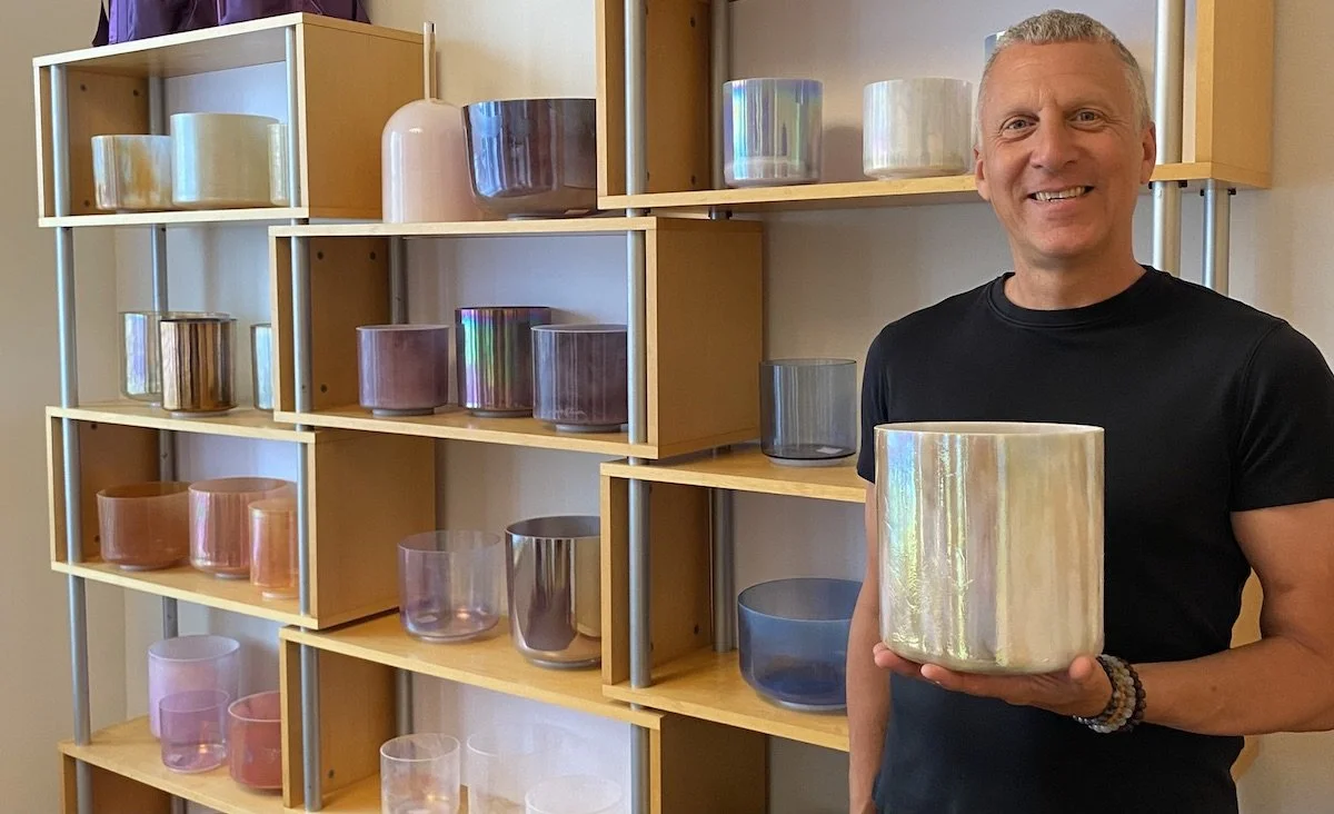 A smiling man in a black t-shirt holding a large iridescent glass piece in front of shelves filled with various colored glass candles or decor items.