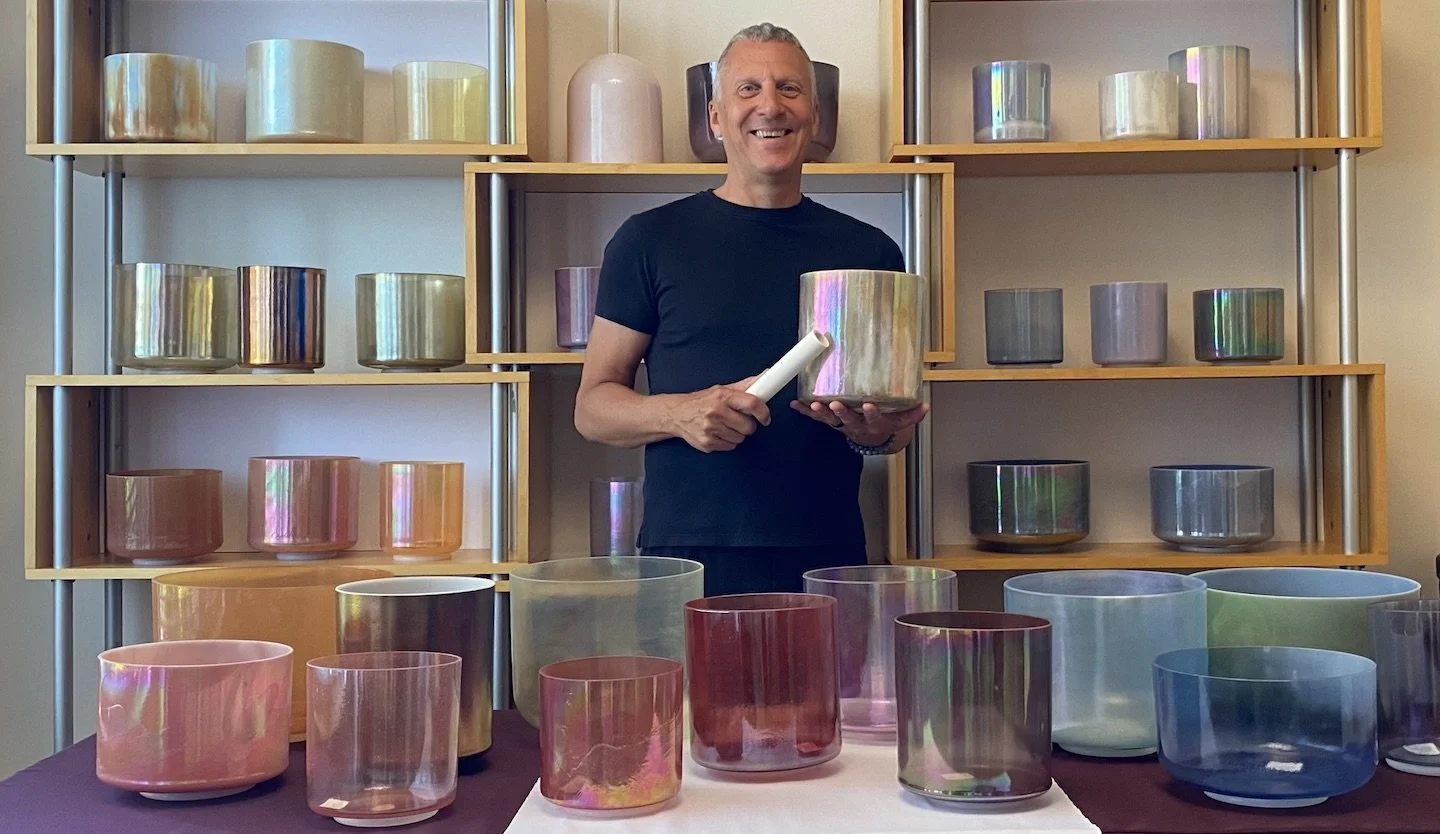 A smiling man holding a tuning fork and a large iridescent bowl in front of shelves filled with similar iridescent bowls of various sizes.