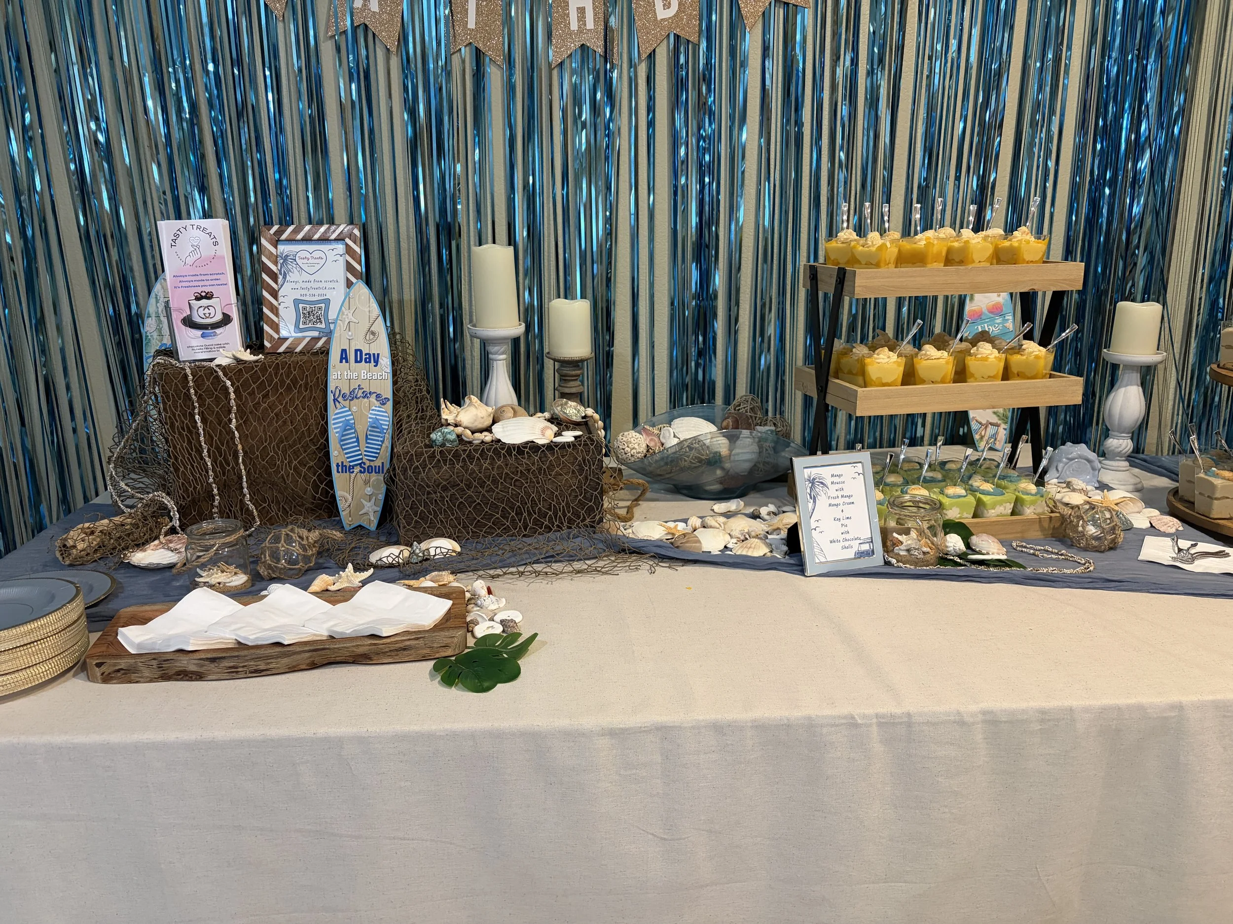Beach-themed dessert table with a shimmering blue backdrop, driftwood signage, and mango mousse dessert cups, on a wooden tiered stand