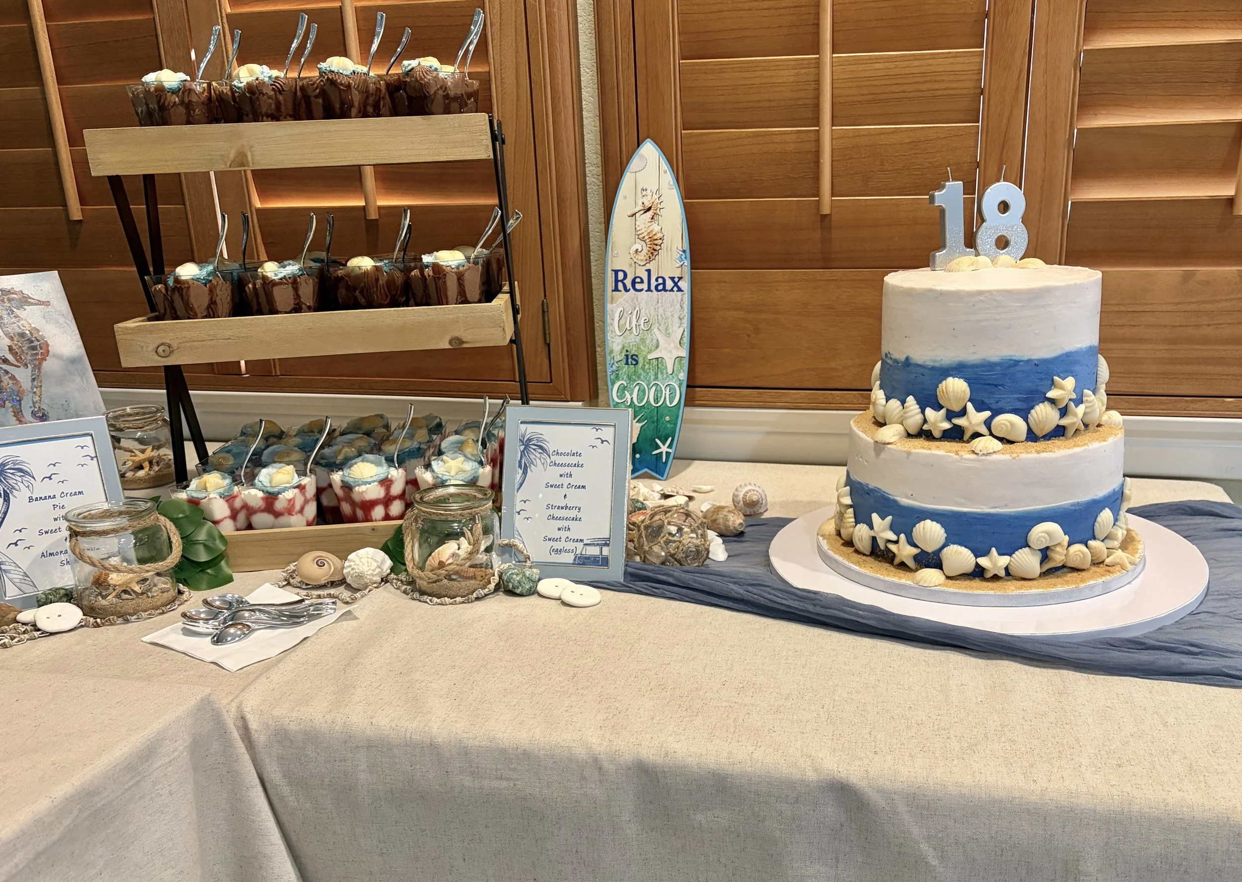 Two-tier blue and white 18th birthday cake with seashell details, displayed beach-themed with a "Relax" surfboard sign
