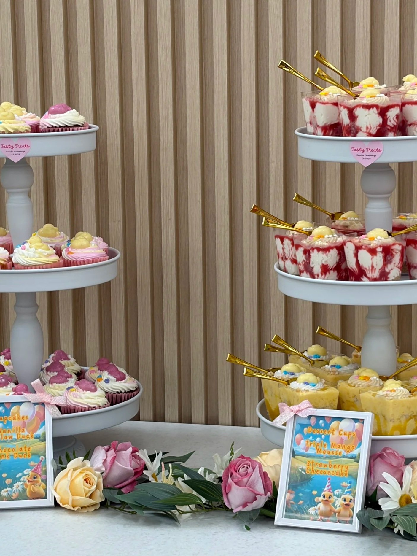 Display of cupcakes and dessert cups arranged on white tiered stands, with decorative flowers and framed signs, set against a wooden slat backdrop.