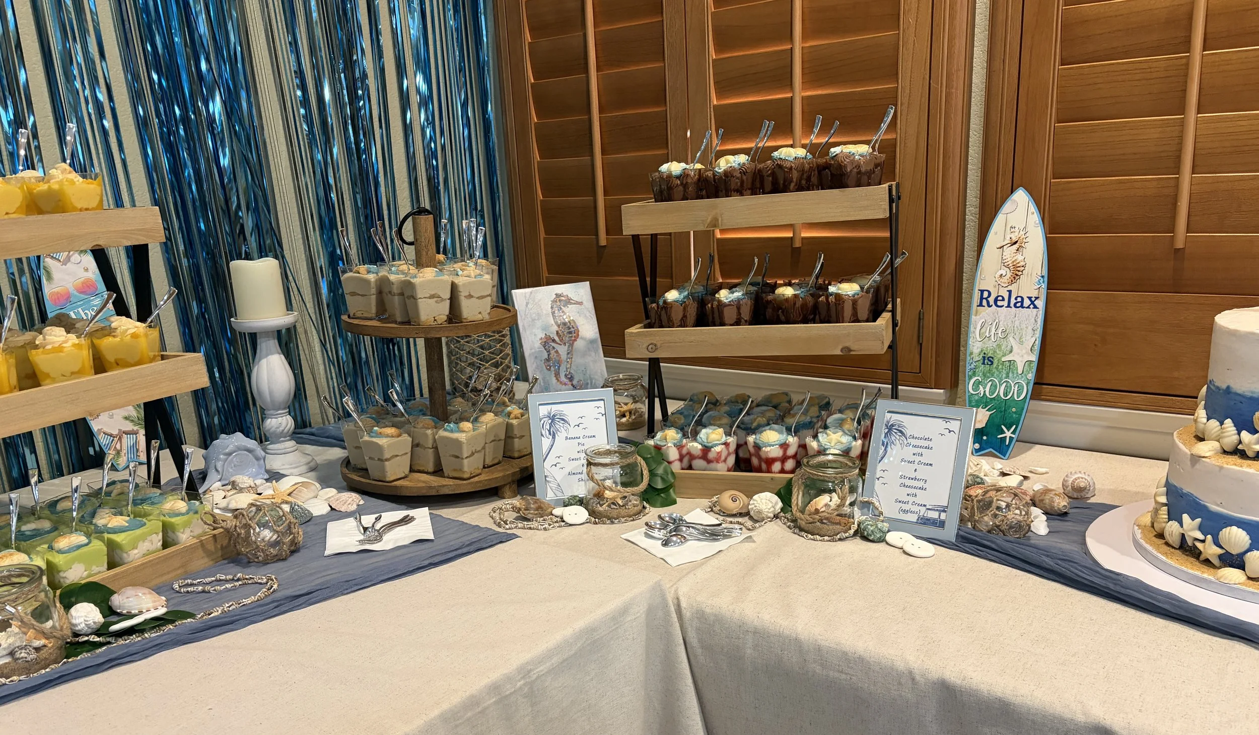 Beach-themed dessert table with a surfboard sign, chocolate cheesecake treat cups, and banana cream pie dessert cups arranged with seashells