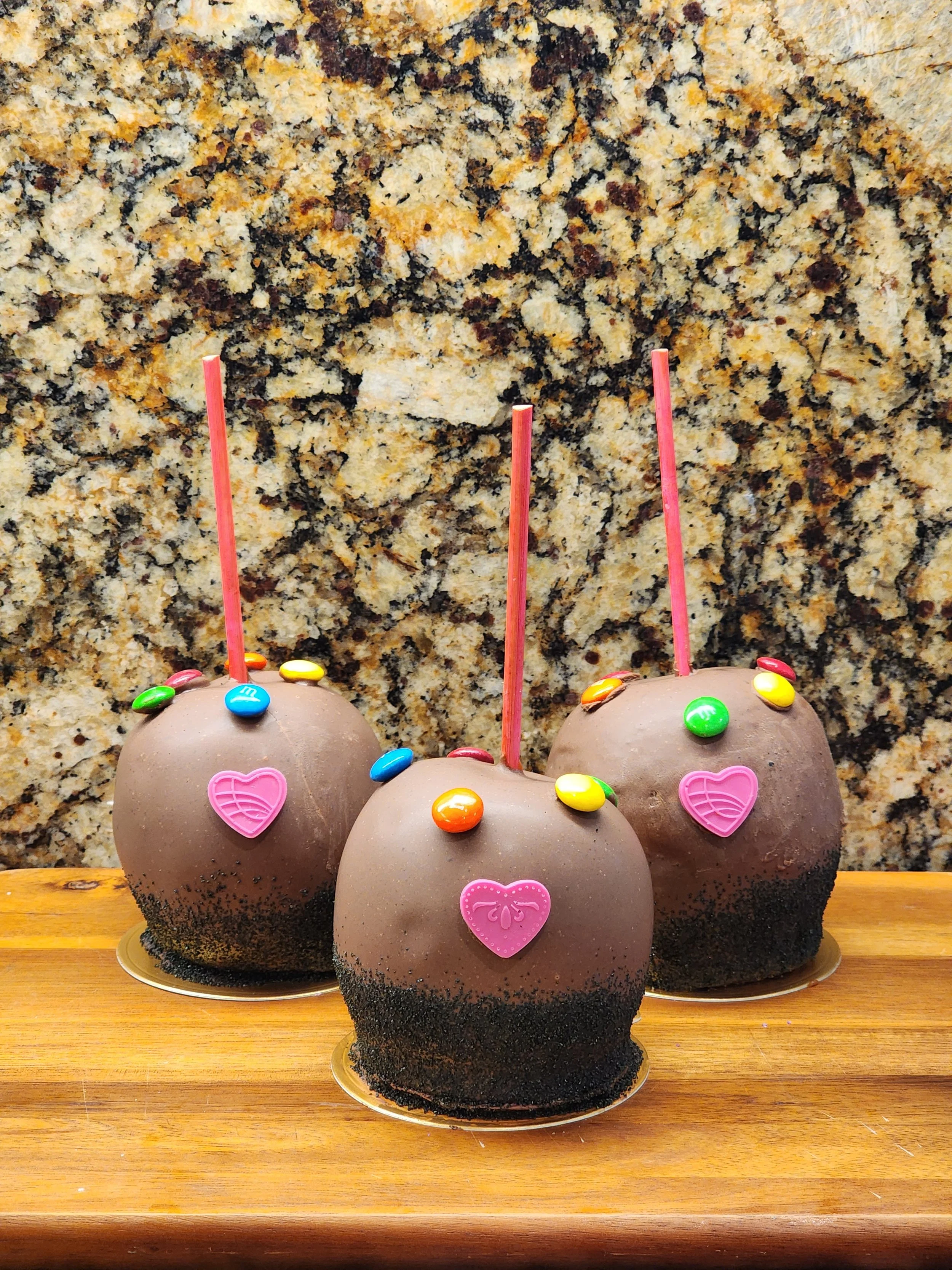Three caramel apples dipped in dark chocolate with pink heart candies and colorful dot candies.