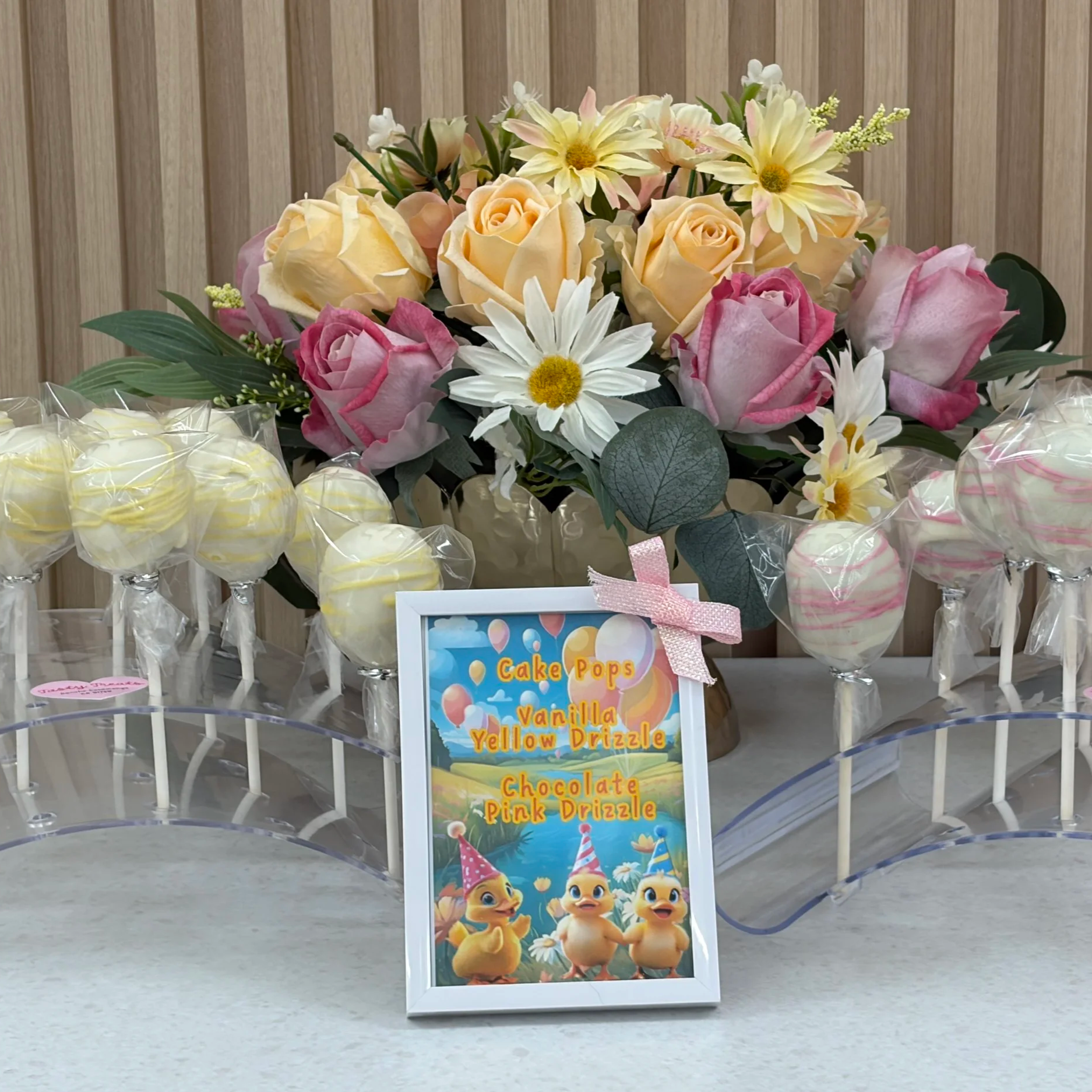 Vanilla and pink-swirl cake pops with a Tasty Treats cake pops sign, displayed among pink and yellow roses