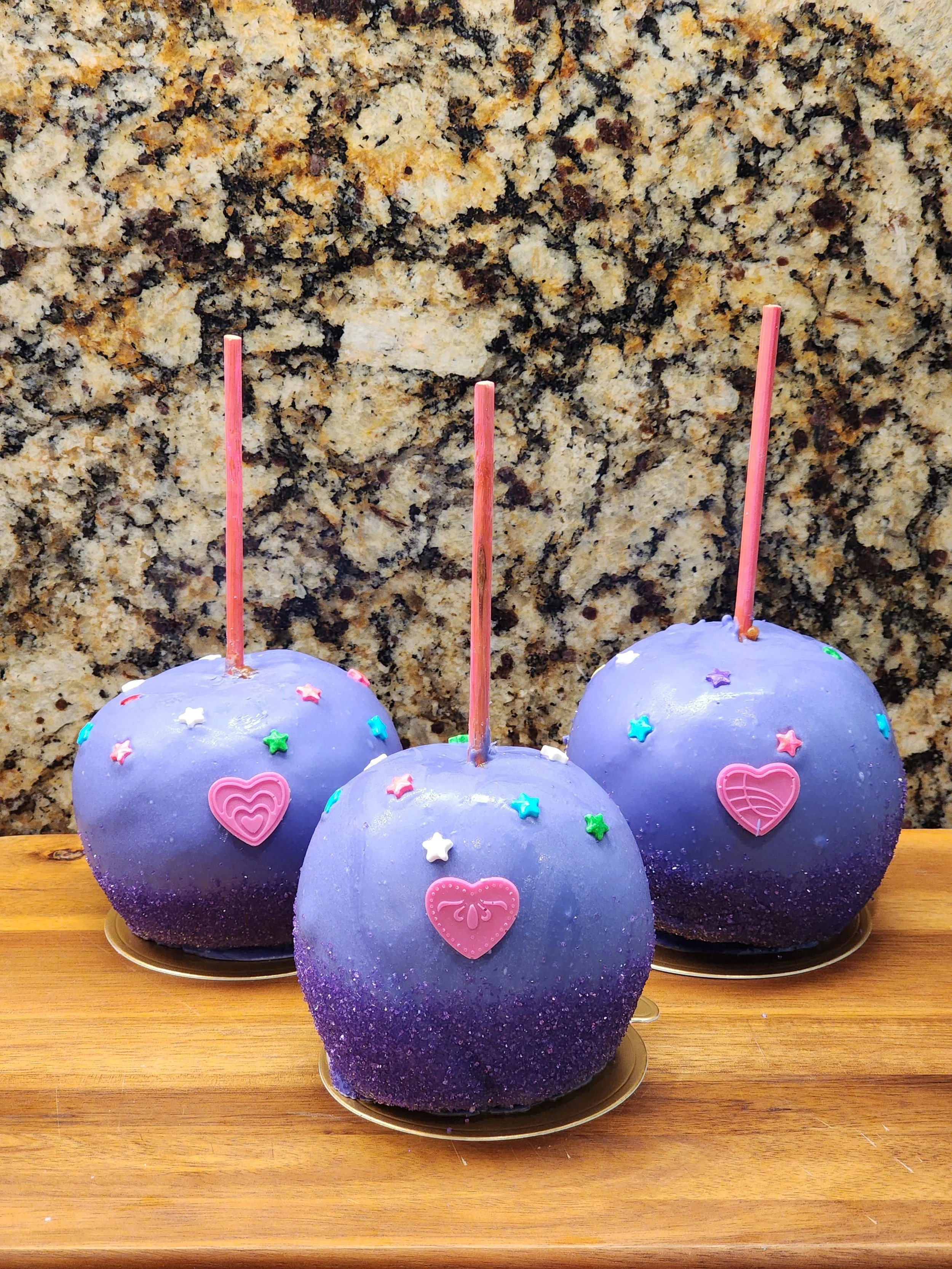 Three caramel apples covered in purple glitter with pink heart candies and star sprinkles.