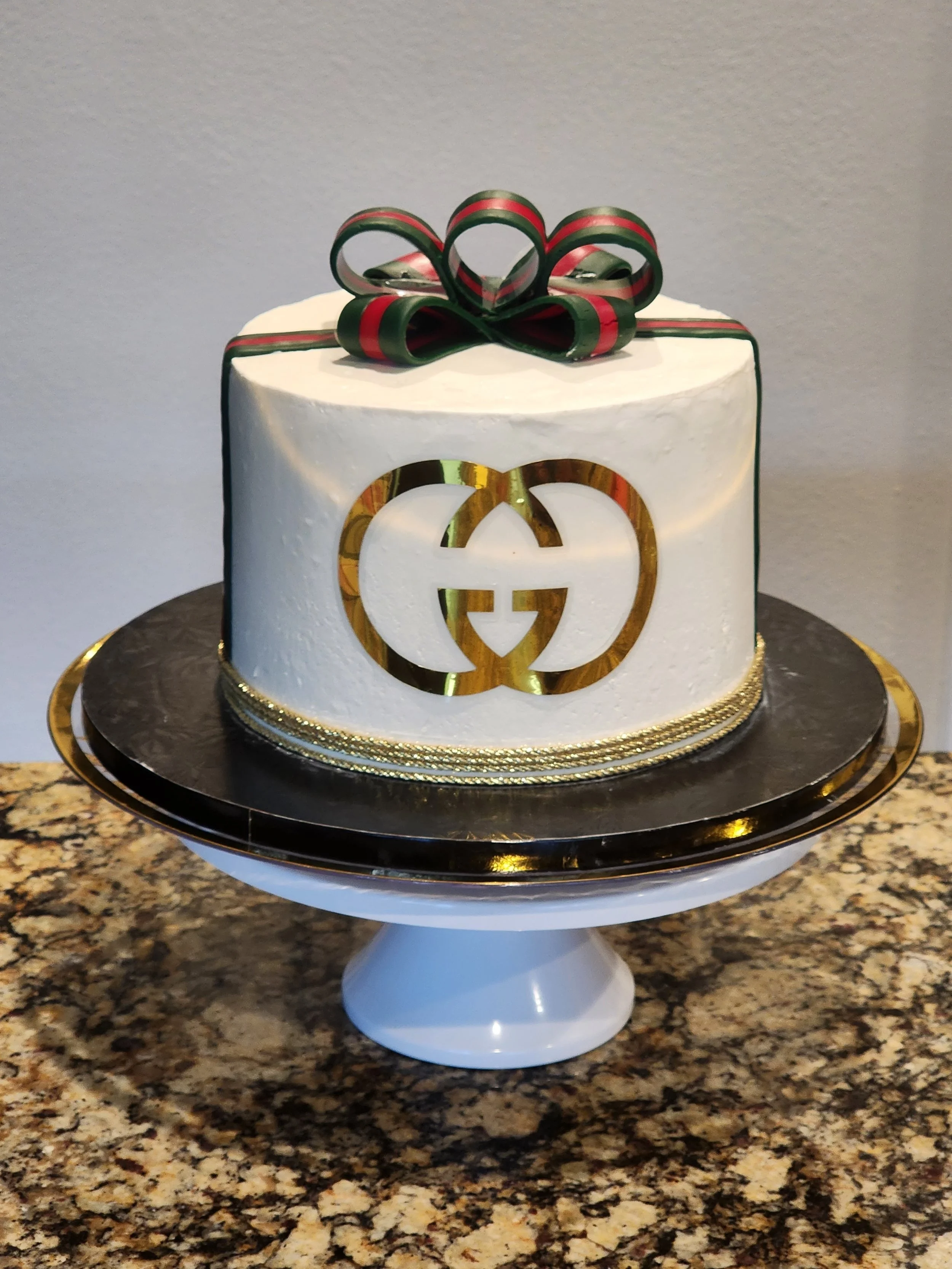 A white cake with a black and gold interlocking logo design and dark rose decorations on top.