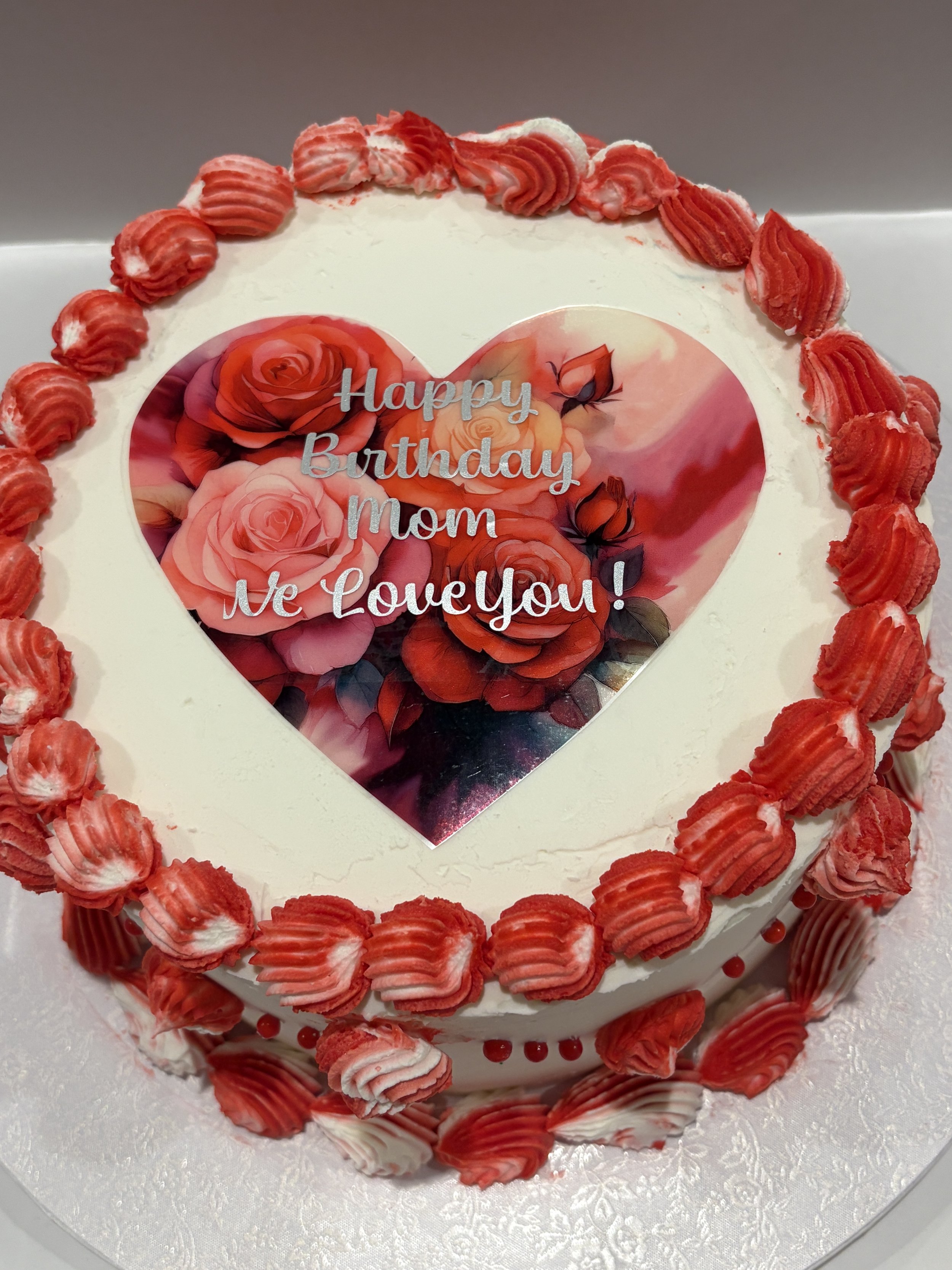 A birthday cake decorated with red and white cream piped rosettes around the edges. The top has an image with a heart shape filled with roses and the message "Happy Birthday Mom We Love You!" written on it.