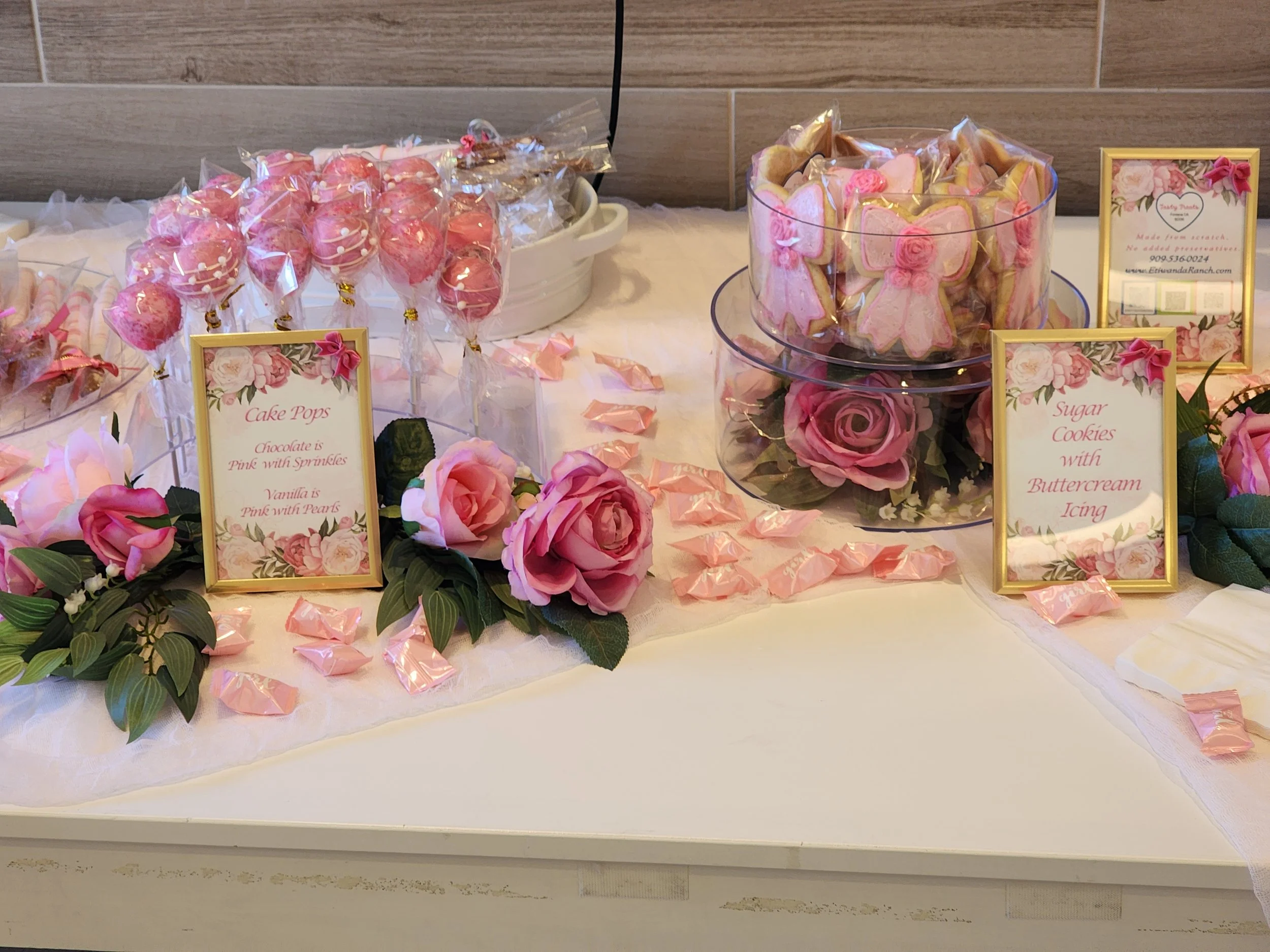 Pink dessert table with wrapped cake pops, bow-shaped sugar cookies, treat cups, and floral arrangements. 