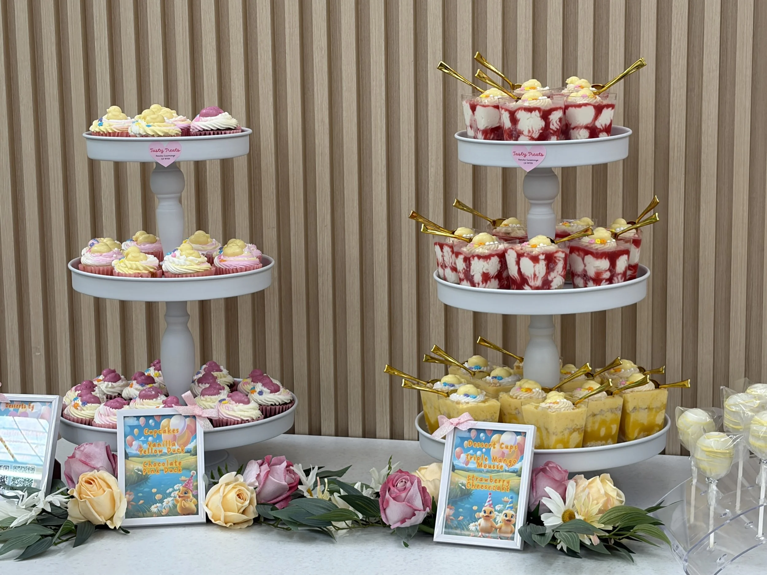 Tiered dessert stand with mini strawberry cheesecakes, mango mousse dessert cup, cake pops, and a mini cake with floral arrangements and mini dessert signs.