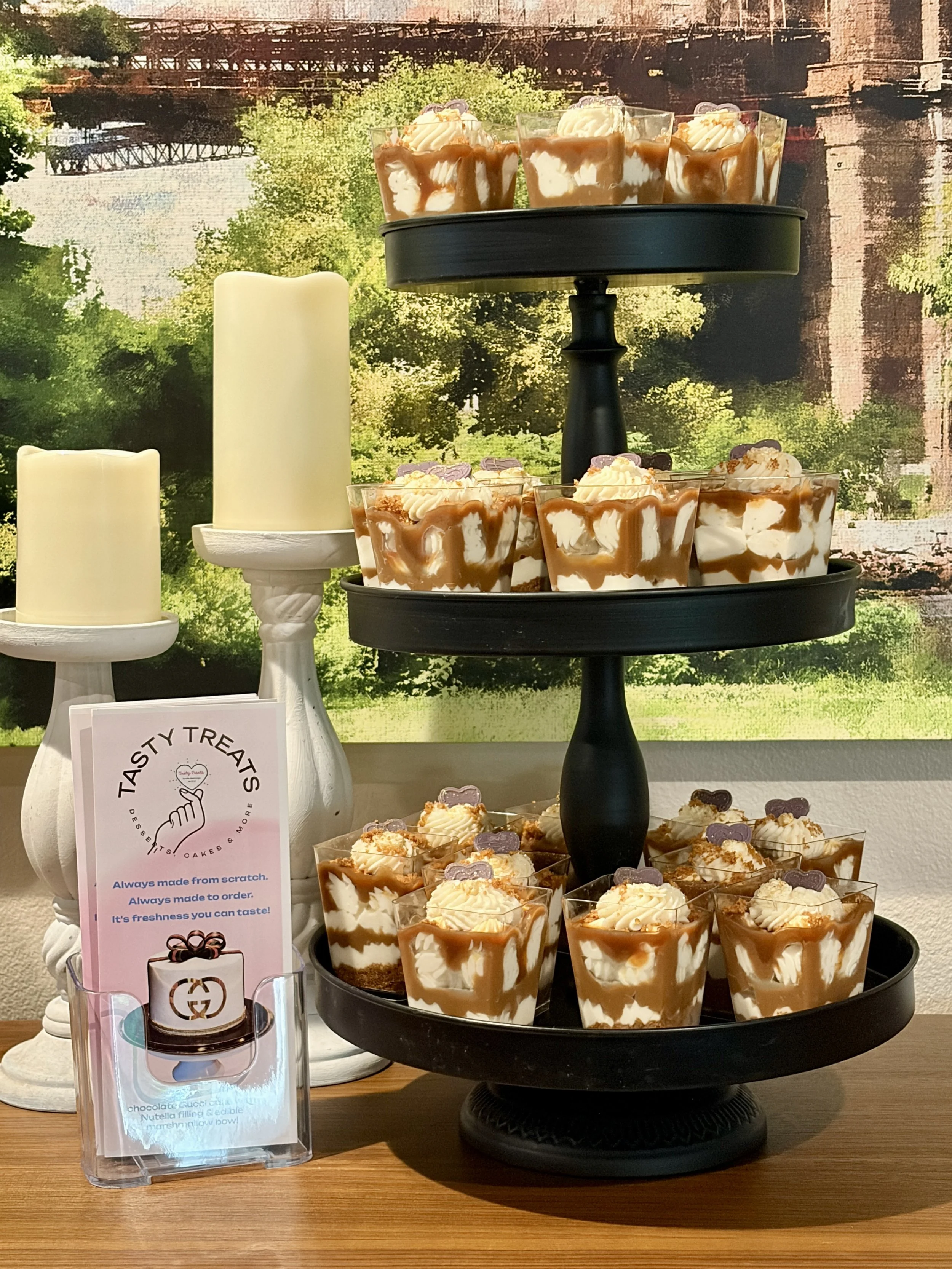 Display of Biscoff cheesecake dessert cups layered with caramel and whipped cream, on a tiered black stand, with two white candles on white stands and a promotional sign for tasty treats.