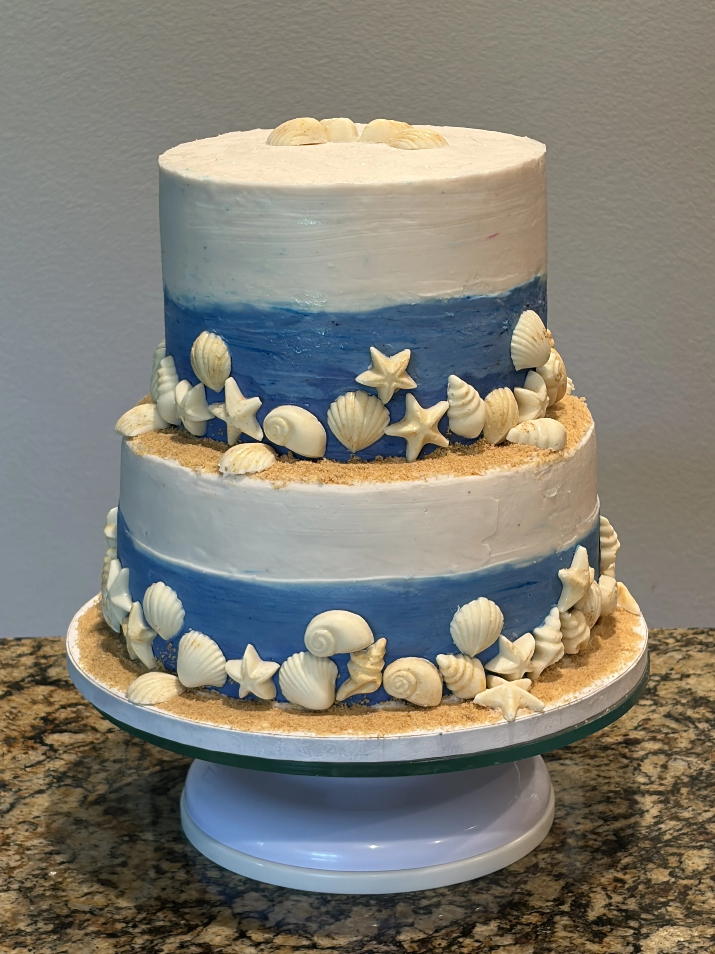 A blue and tan two-tier cake decorated with seashells in a beach theme.