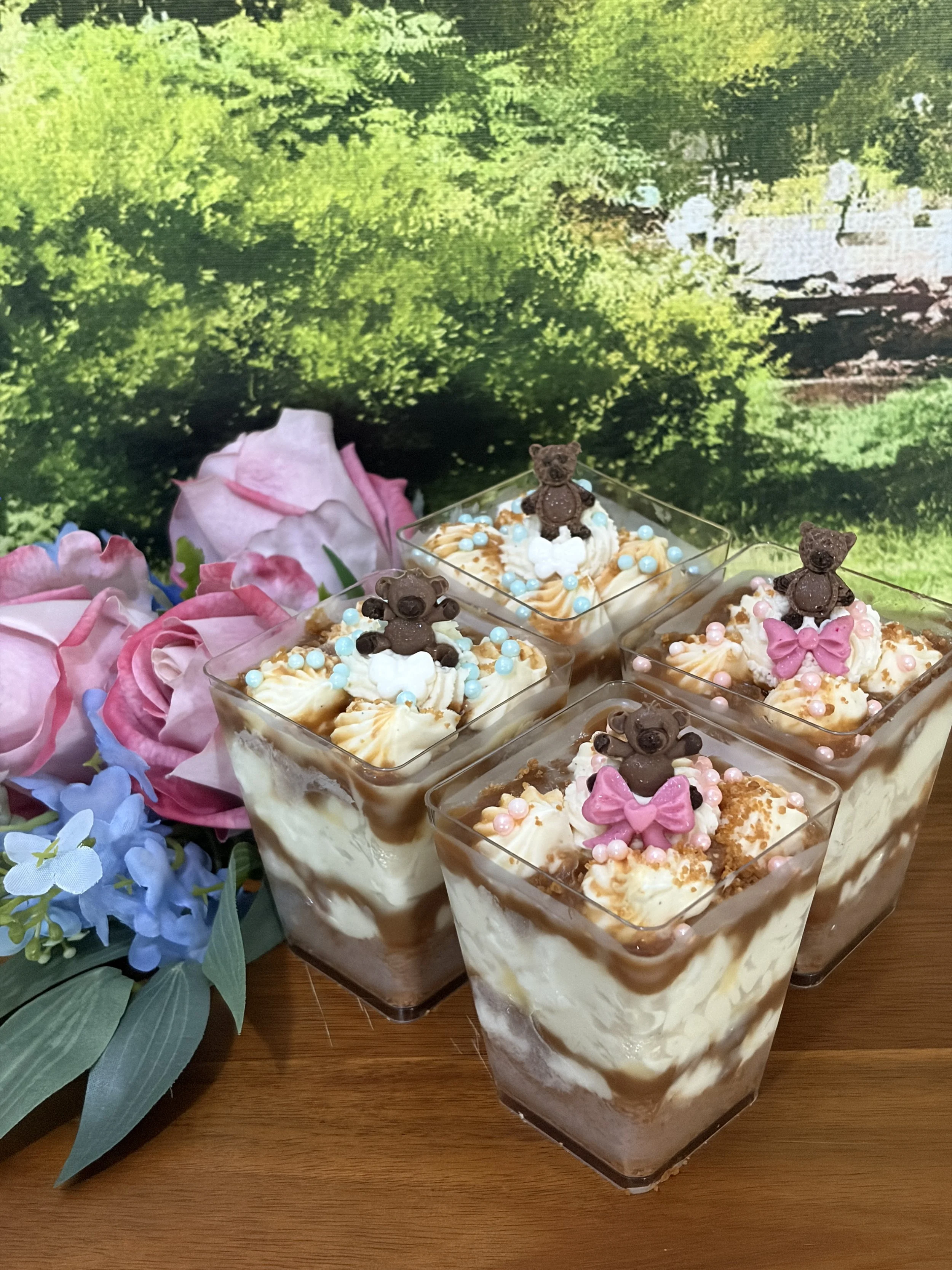 Biscoff cheesecake dessert cups topped with whipped cream and mini chocolate teddy bears, on a wooden serving tray and a floral arrangements. 
