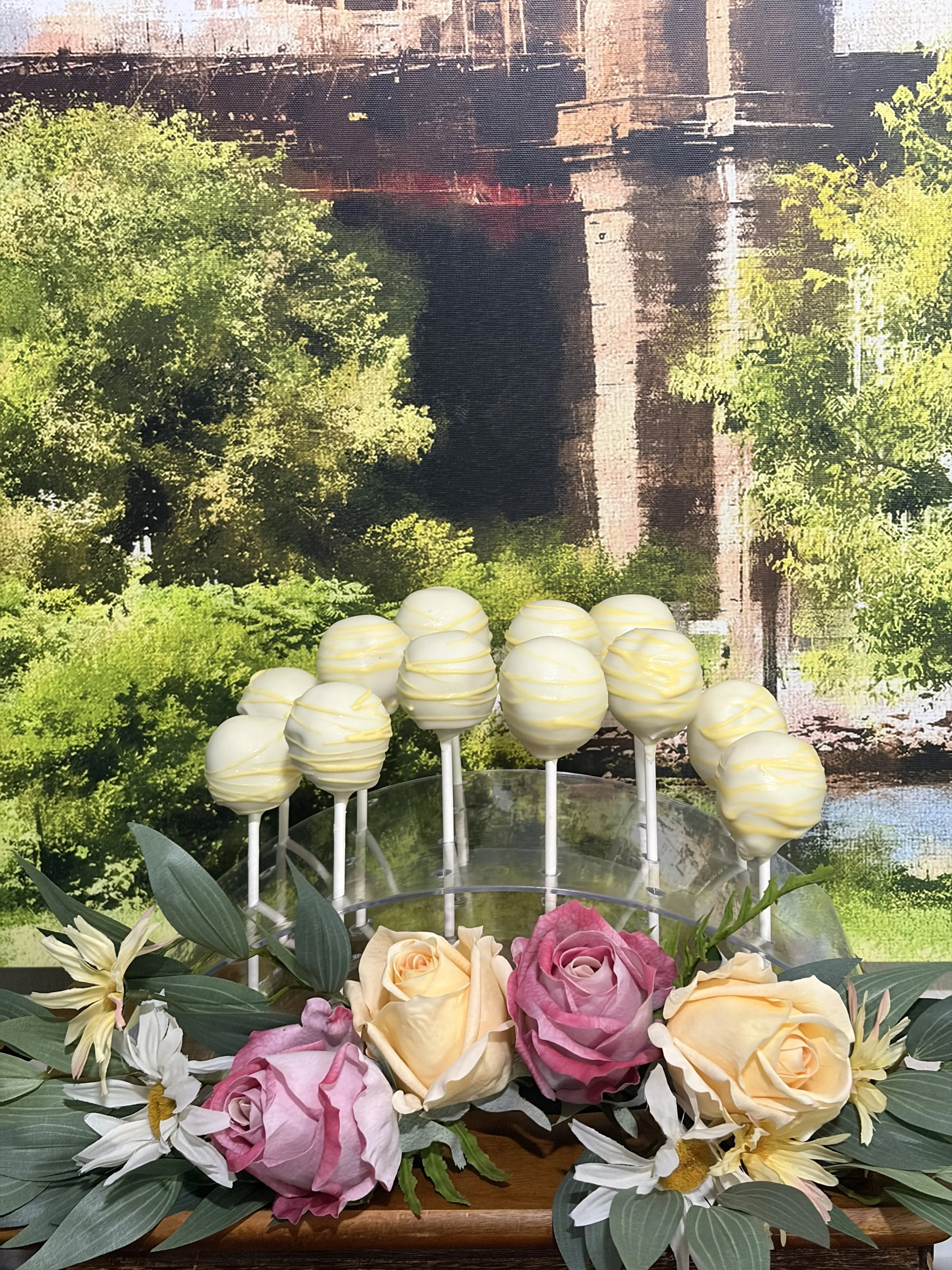 Vanilla yellow-drizzle and pink-swirl cake pops displayed with a Tasty Treats cake pops sign, surrounded by pink and yellow roses