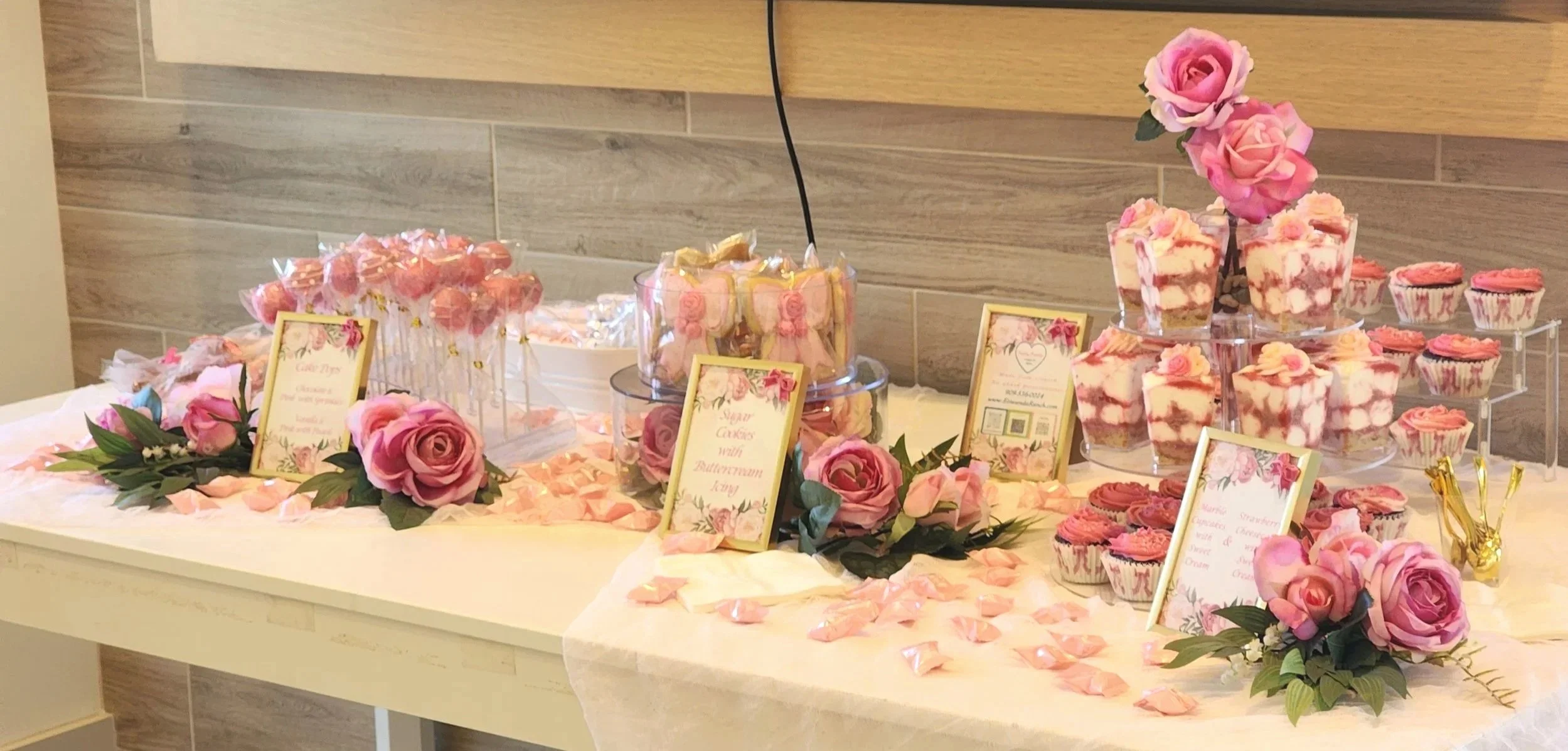 Pink-themed dessert table with strawberry cheesecake dessert cups, mini dessert signs, and scattered rose petals