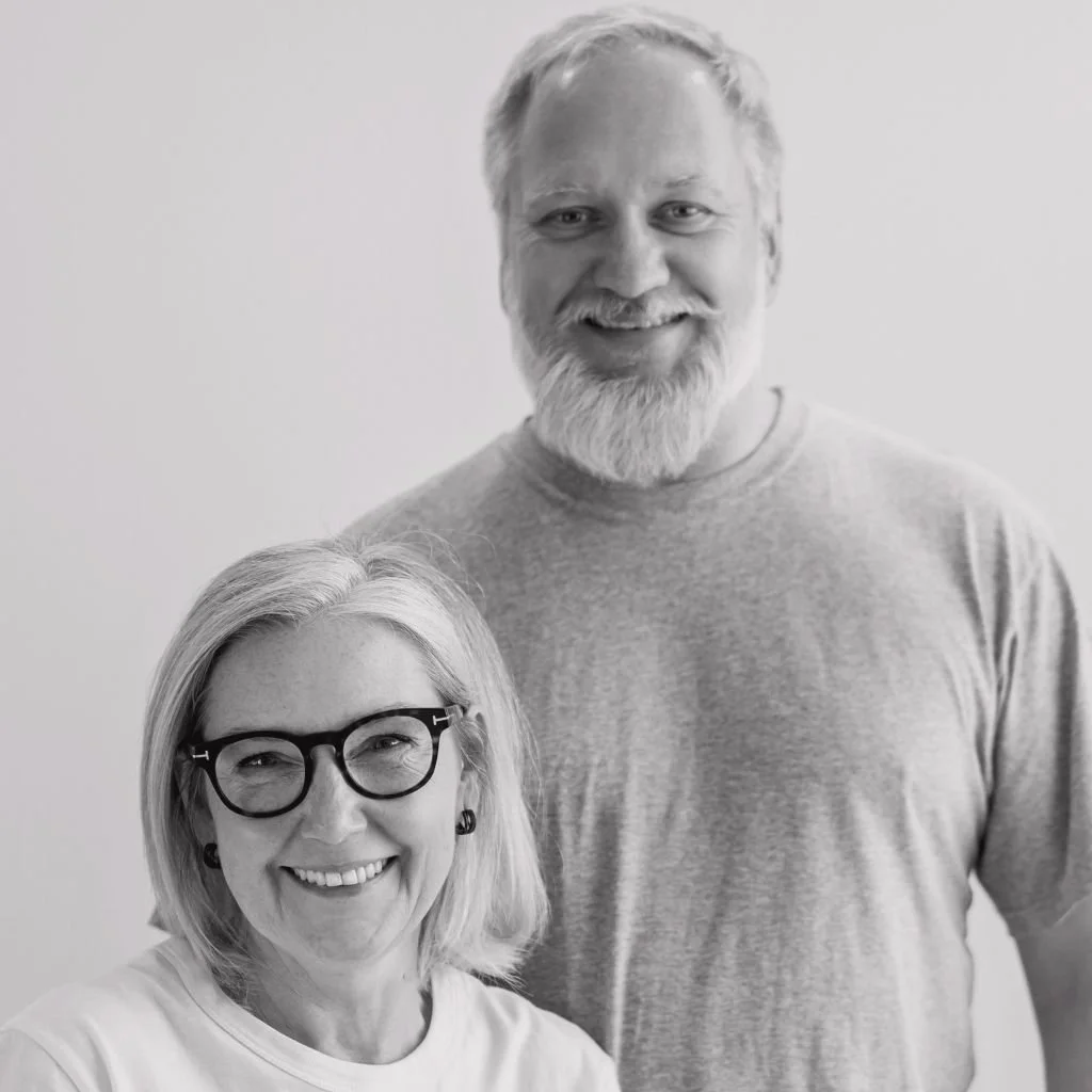 A black-and-white photograph of a smiling man with light hair, a beard, and glasses, and a smiling woman with medium-length hair, glasses, and earrings, standing against a plain light background.