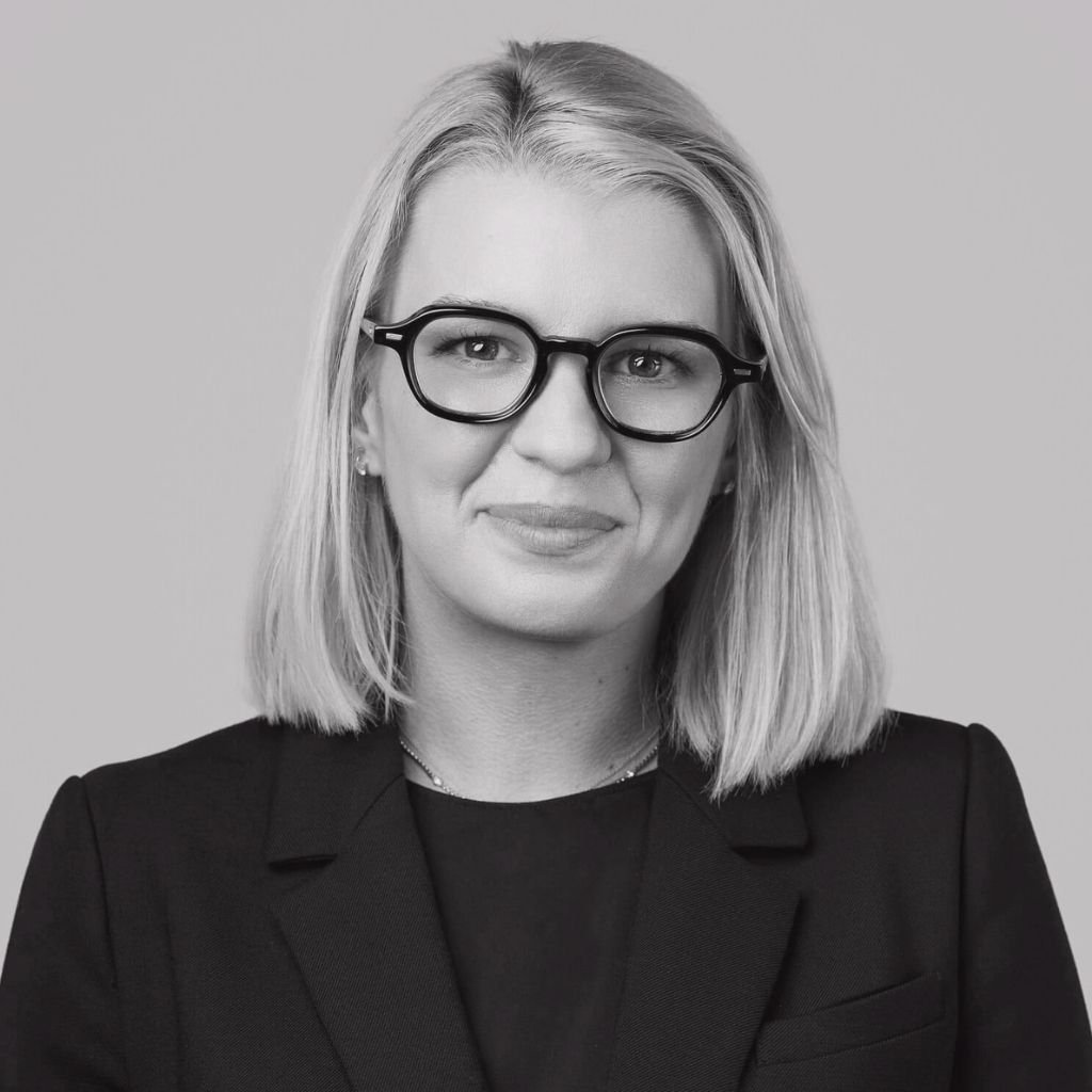 Black and white portrait of a woman with shoulder-length blonde hair wearing glasses and a dark blazer, against a plain background.
