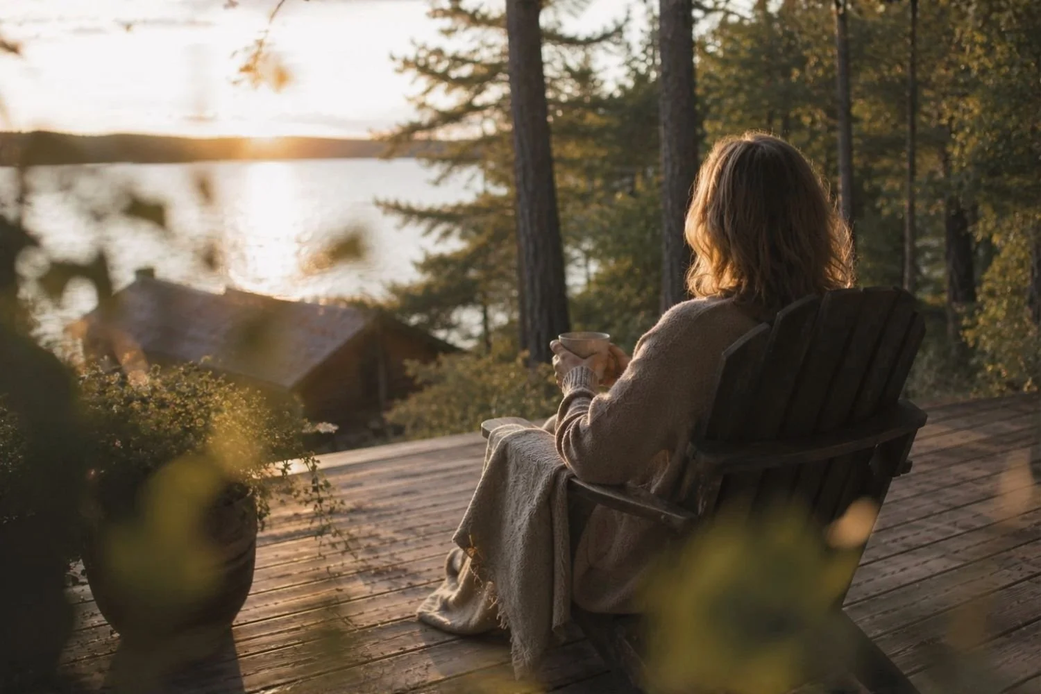 Rear view of a woman enjoying a quiet moment in a wooden deck chair, looking out over a calm lake at sunset during retirement.