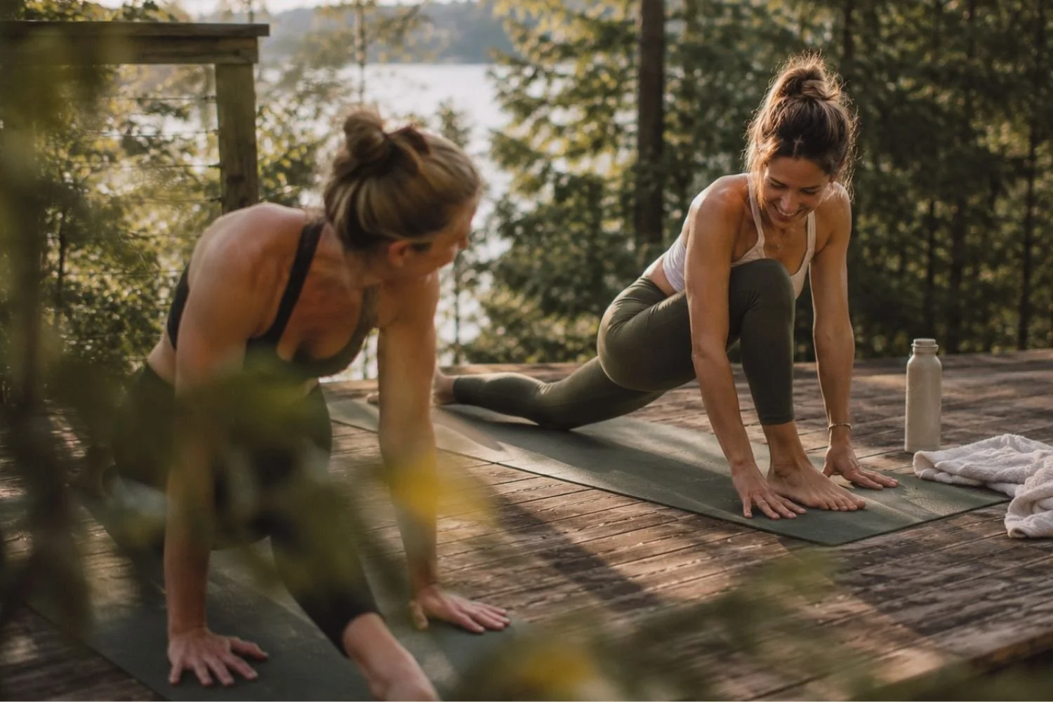 Two women practicing yoga by a peaceful lake in warm light, highlighting how managing money and mental health requires slowing down.