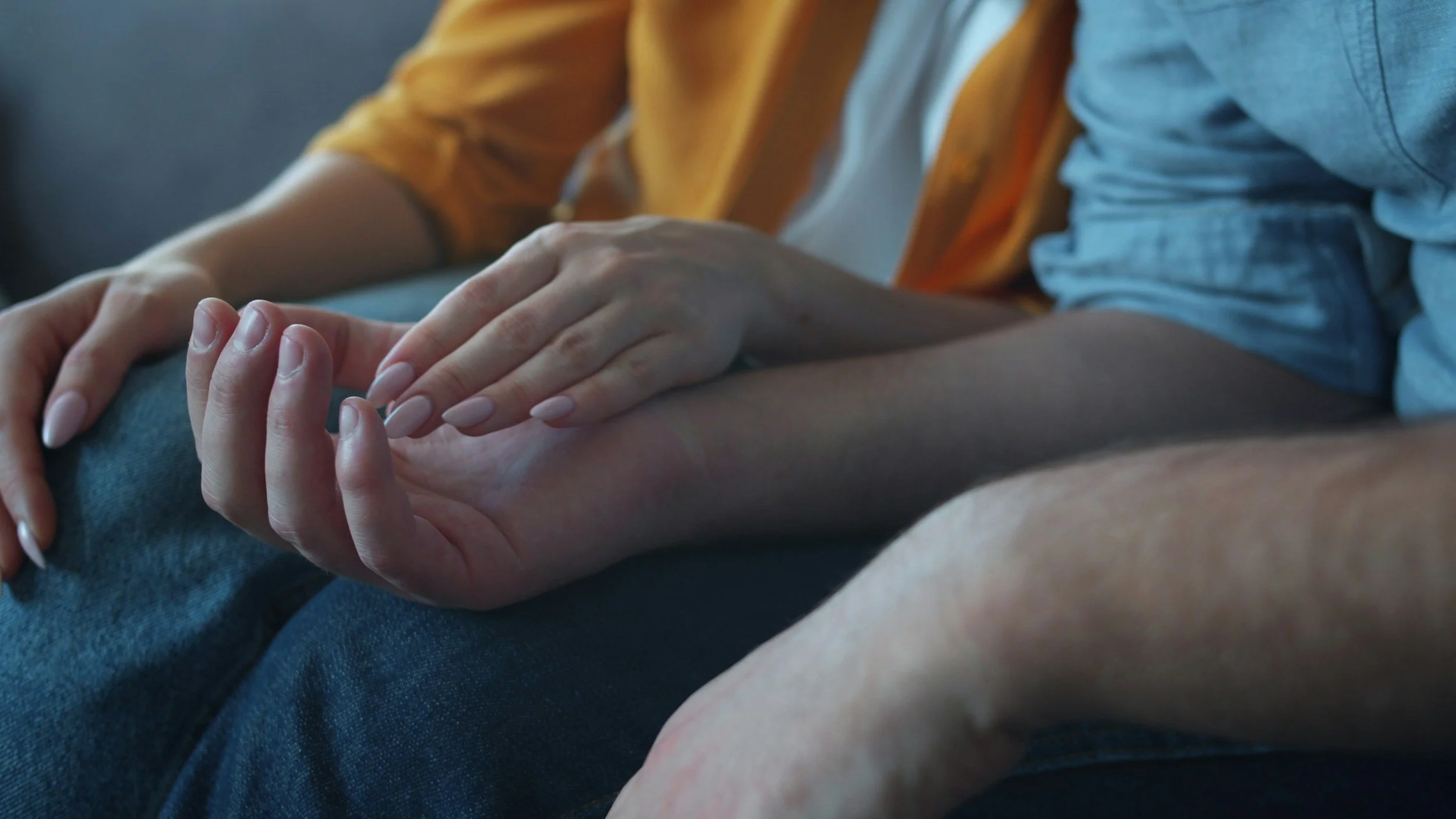 Close-up of two people holding hands, one with a light-colored manicure and the other with a darker hand, sitting close together.
