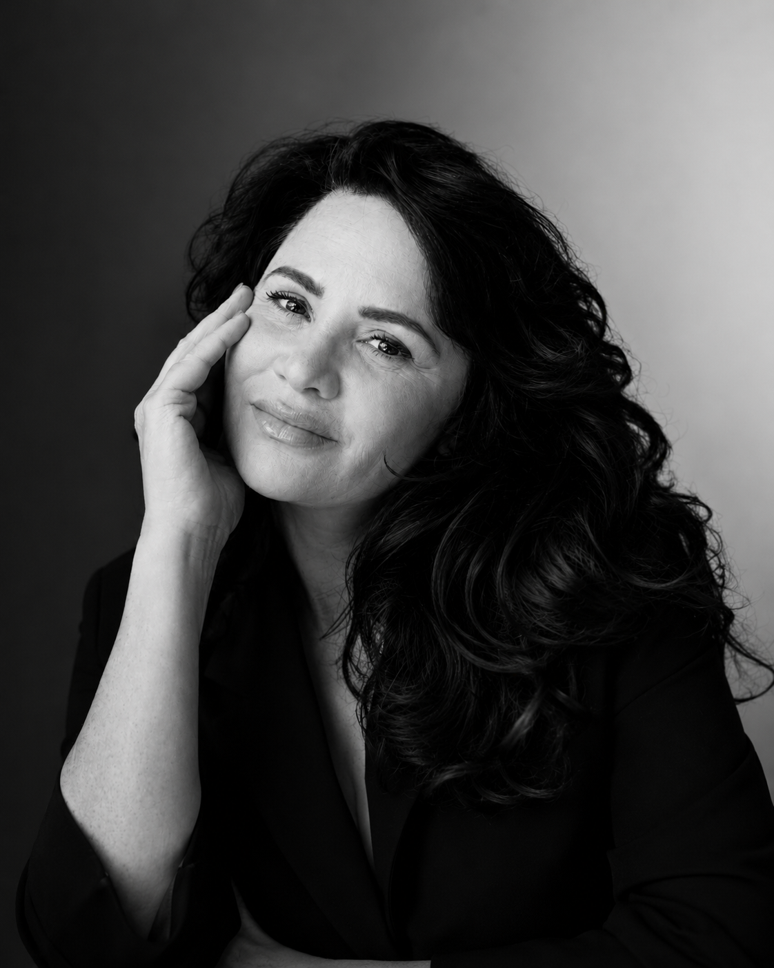 Black and white portrait of a woman with long, wavy hair, resting her head on her hand and smiling softly.