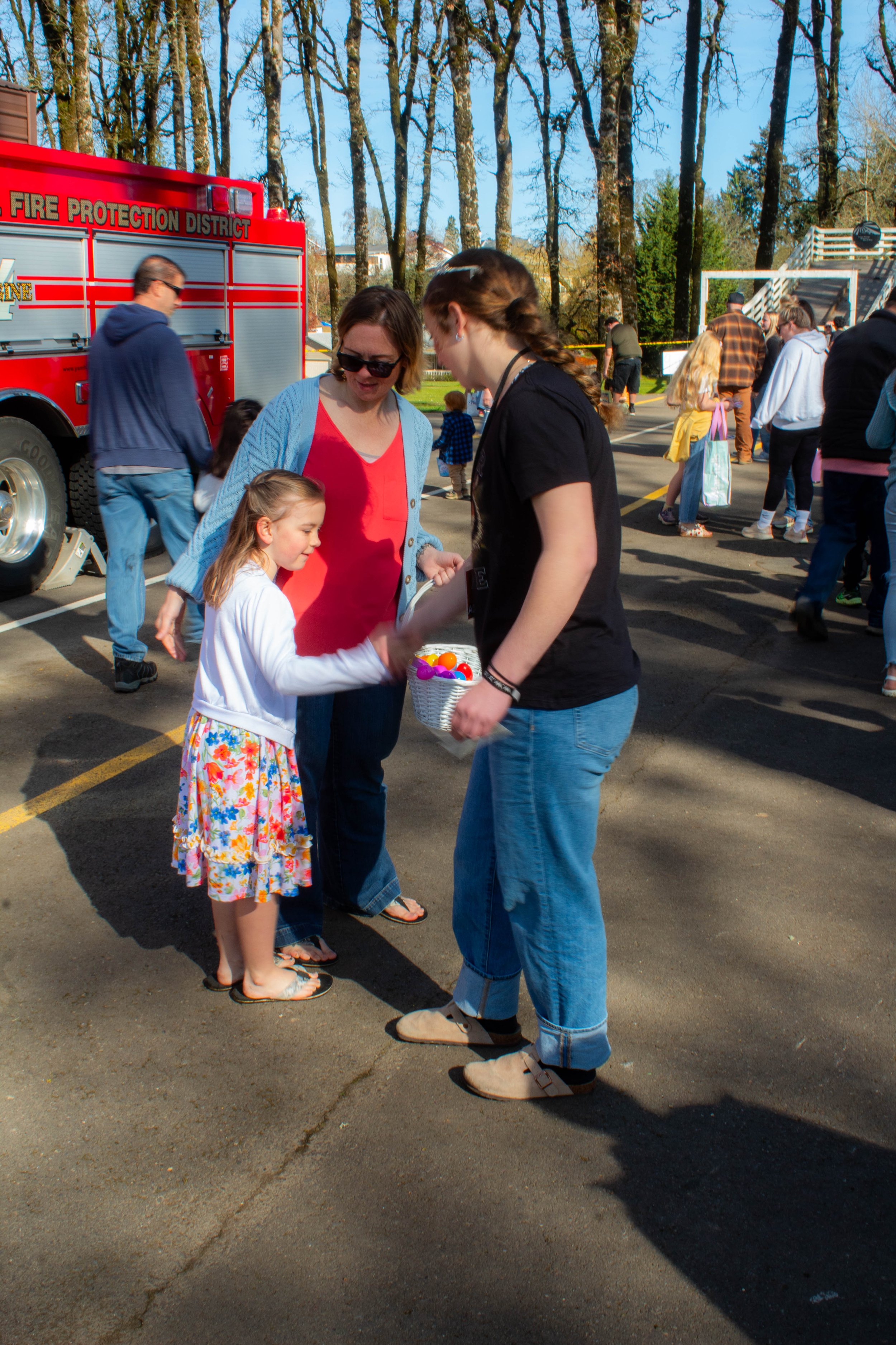 A young girl in a floral skirt and white jacket receives a small toy or prize from a woman in a black t-shirt at an outdoor event. An older woman in sunglasses looks on, while a fire truck is visible in the background among other people at a park or community gathering.