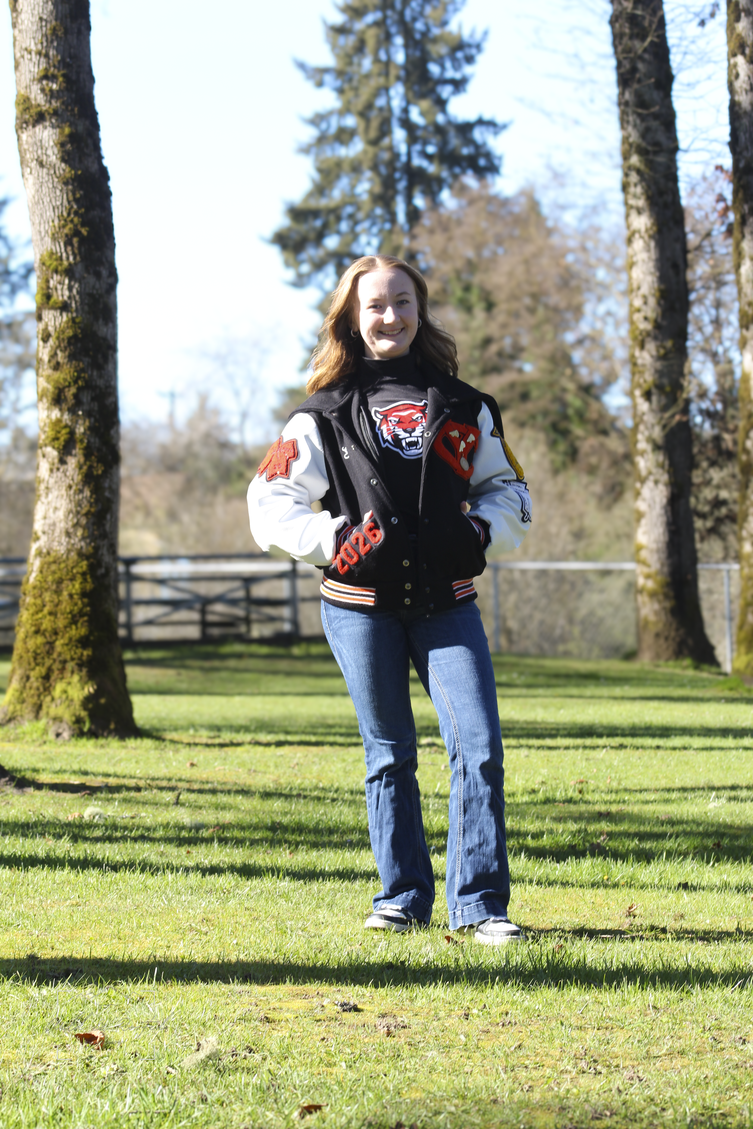 A young woman with red hair standing outdoors on a grassy area, smiling, wearing a black and white varsity jacket, jeans, and sneakers, with trees and a fence in the background.