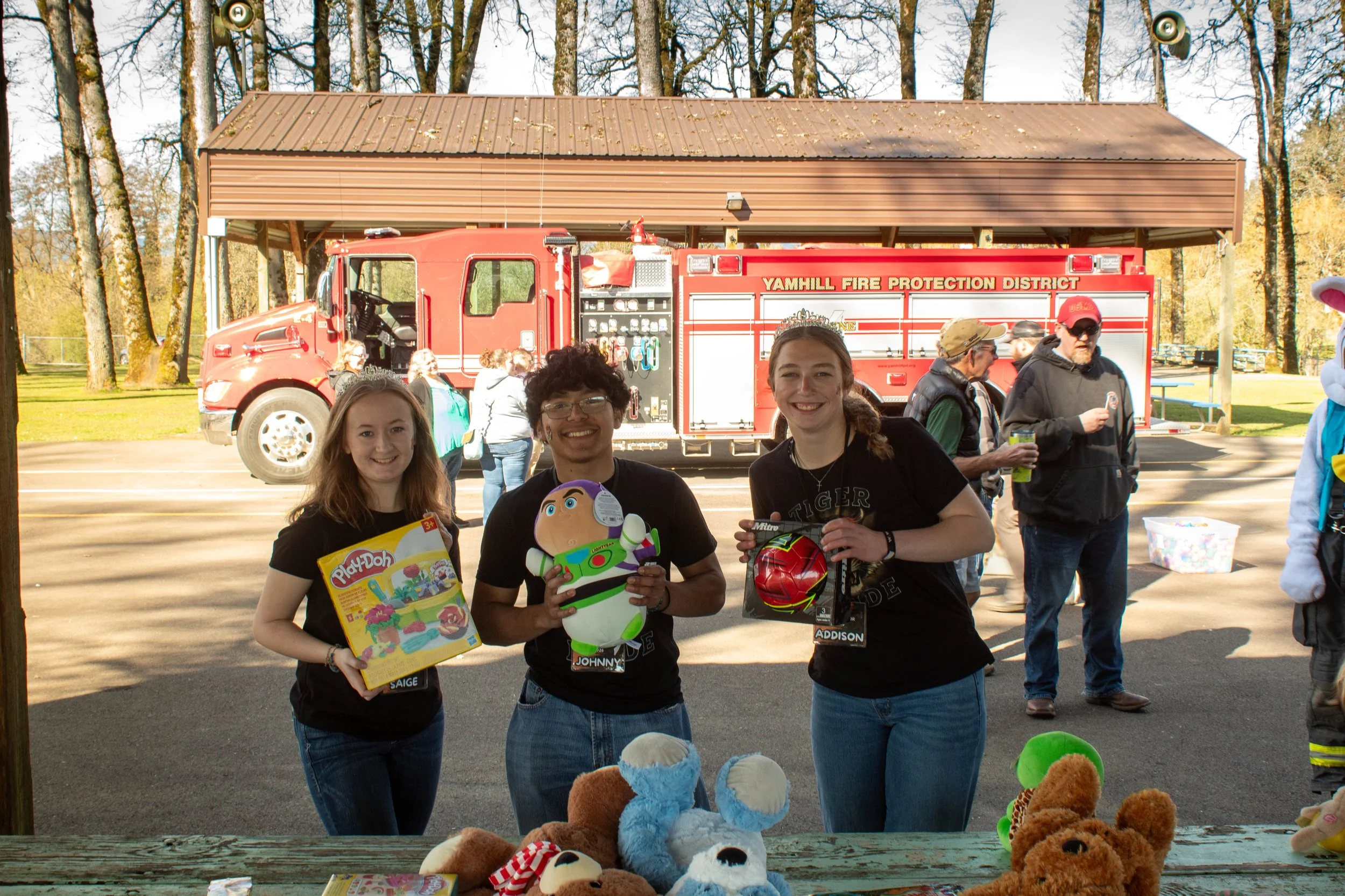 Three young women standing behind a table with plush toys, holding toys and smiling at the camera. Behind them is a fire truck and other people at an outdoor event under a shelter with trees in the background.