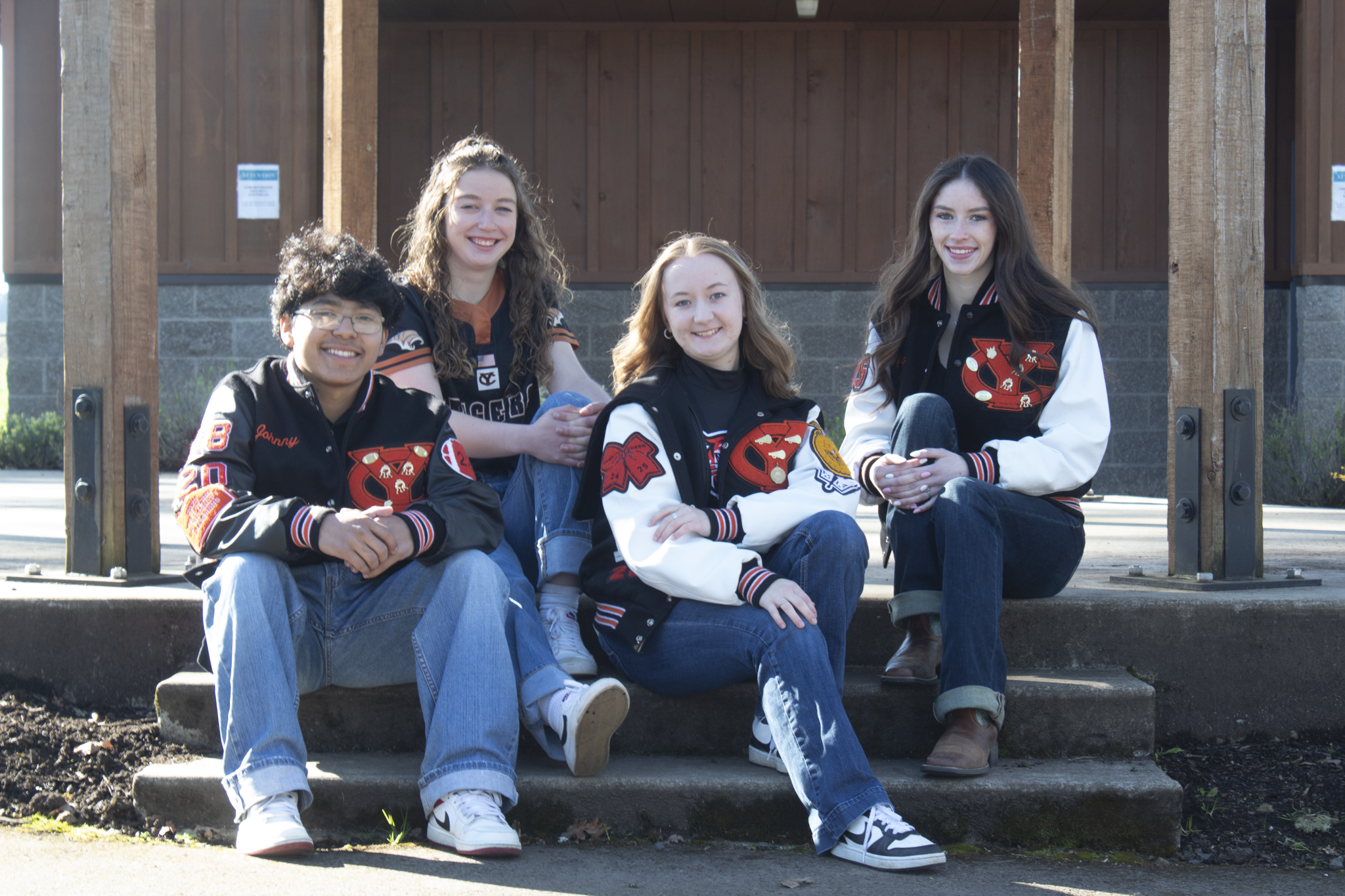 Five teenage girls sitting and kneeling on outdoor stairs, wearing varsity jackets with patches and smiling at the camera.