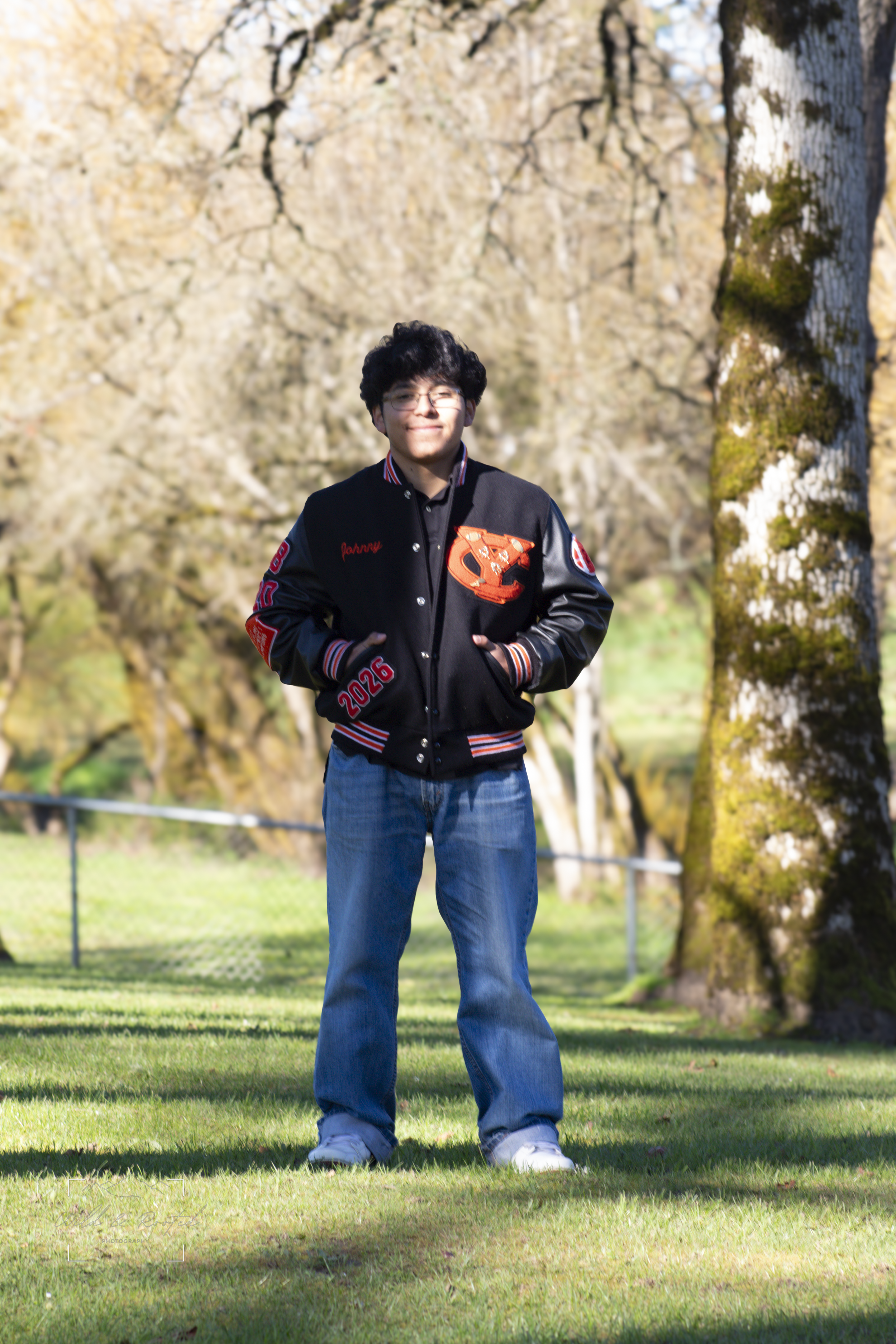 A young man standing outdoors on a grassy area, surrounded by trees, wearing a black and orange varsity jacket with patches and the year 2026, blue jeans, and white sneakers, smiling at the camera.