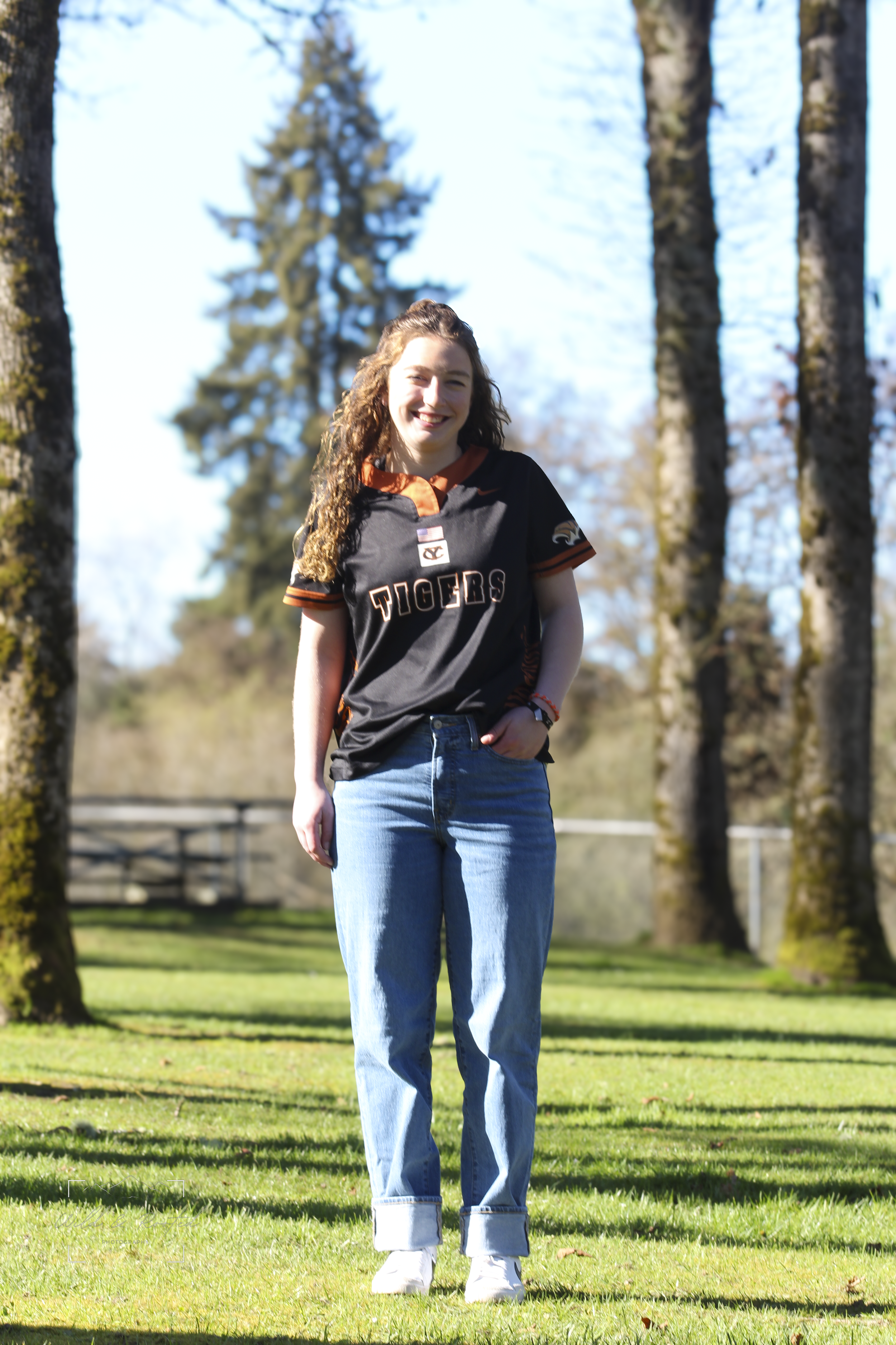 A young woman with long curly hair standing outdoors on a sunny day, wearing a black sports jersey with 'TIGERS' written on it, blue jeans, and white shoes, smiling at the camera with trees and a grassy field in the background.