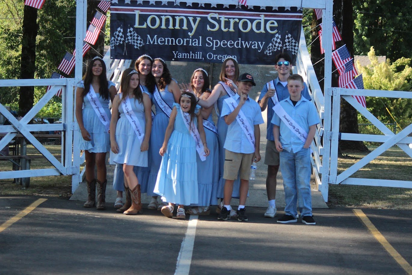 A group of children and teens dressed in light blue and white, standing on a staircase at the Lonny Strode Memorial Speedway in Yamhill, Oregon, celebrating with sashes for a parade or event, decorated with American flags.