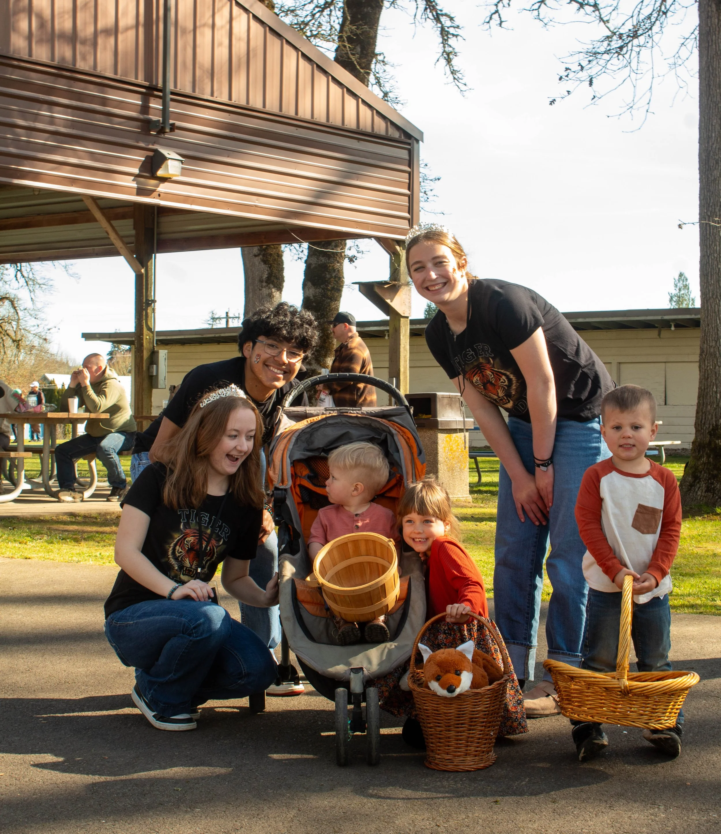 A group of six people, including children and young adults, posing together outdoors in a park-like setting with picnic tables and trees, some holding baskets, with a stroller and plush animal, smiling and enjoying a sunny day.