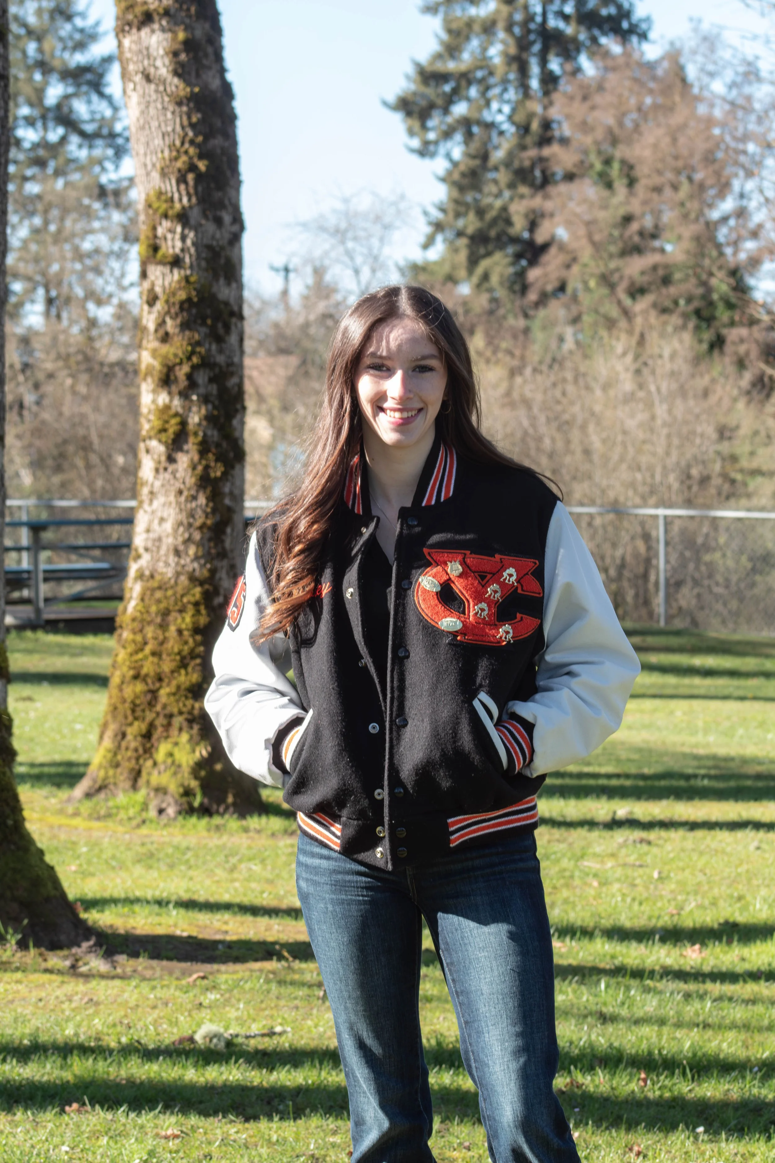 A young woman with long brown hair smiling outdoors, wearing a varsity jacket with letter patches, blue jeans, and standing on a grassy area with trees in the background.